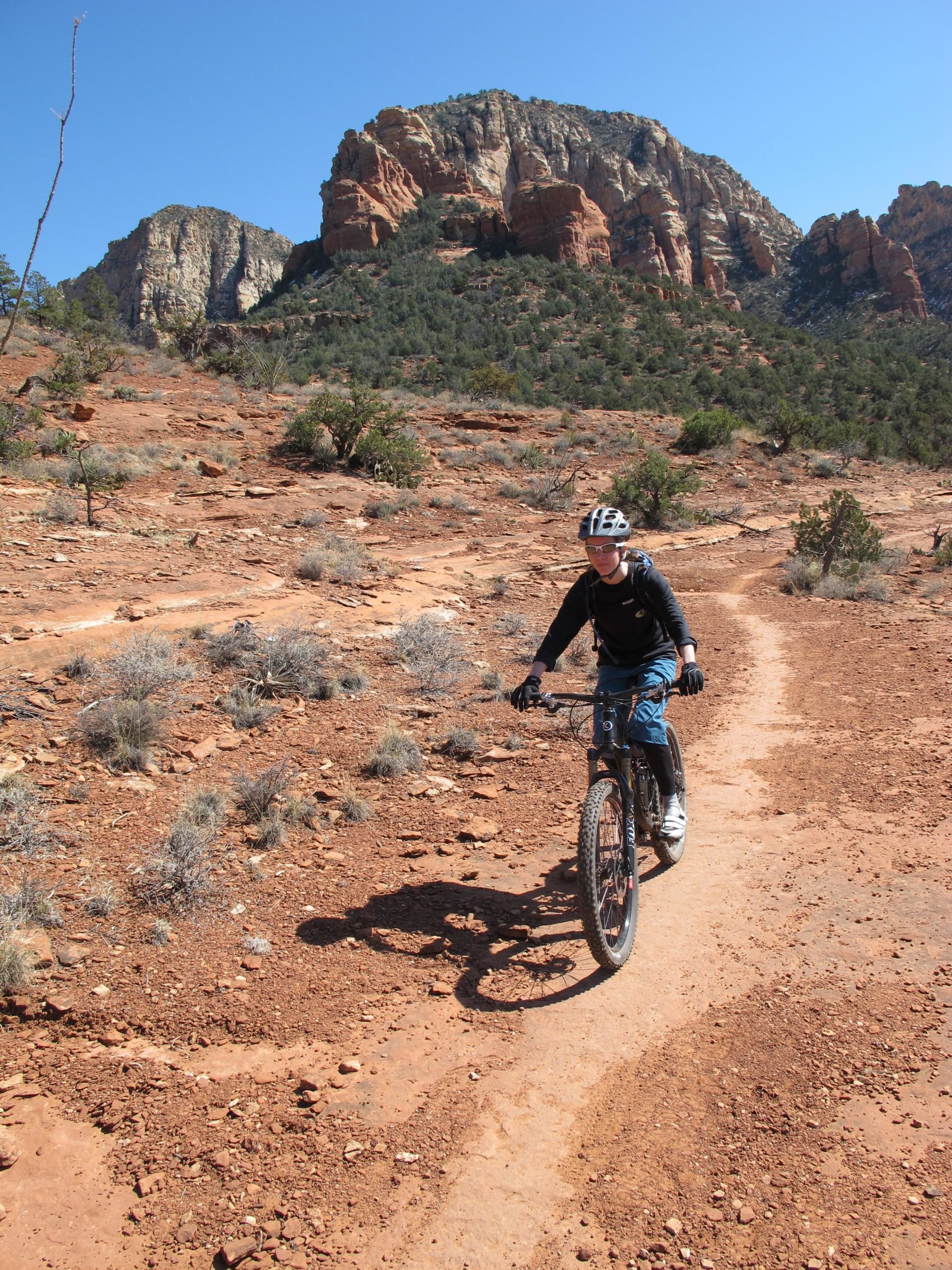 A person riding a mountain bike along a dirt trail in a rocky desert landscape, with large rock formations and blue sky in the background. Bell Rock Area Trails mountain bike trail.
