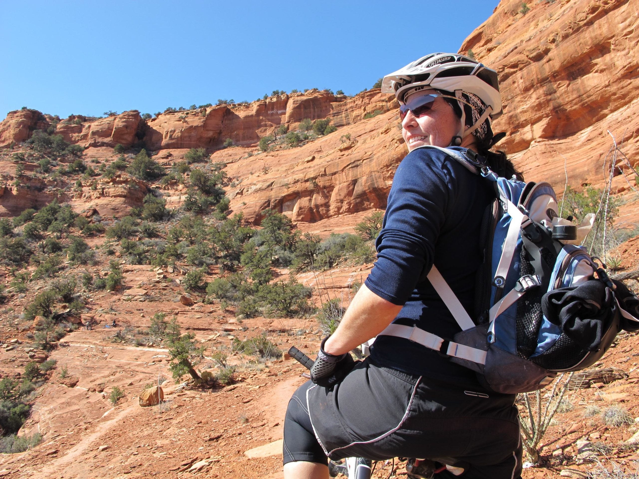 A mountain biker wearing a helmet and sunglasses rests on their bike, looking back at the rocky landscape filled with red cliffs and greenery under a clear blue sky. Bell Rock Area Trails mountain bike trail.