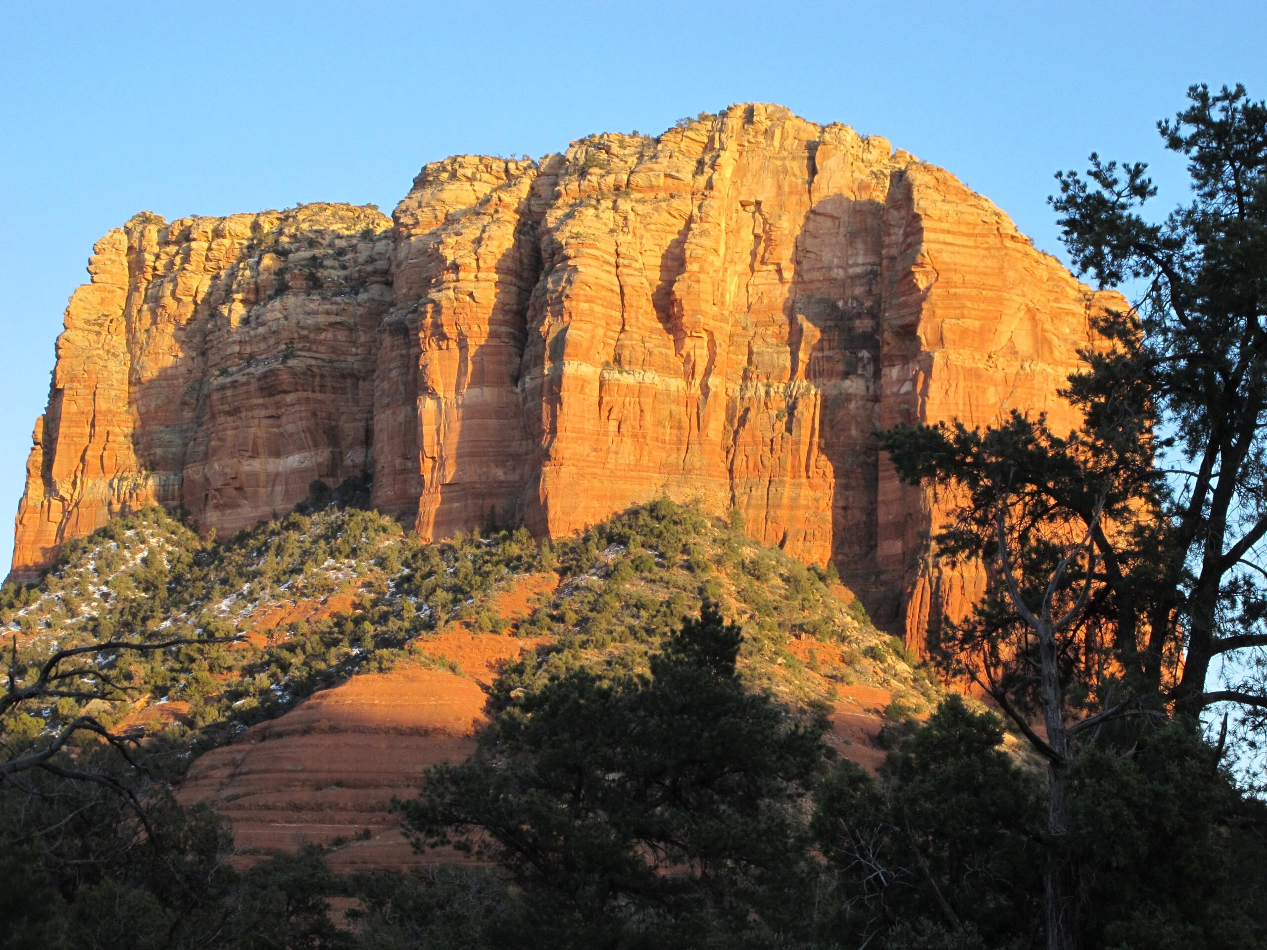 A towering red rock formation illuminated by the setting sun, surrounded by green vegetation and trees under a clear blue sky. The rugged texture of the cliffs is highlighted by shadows and layers, showcasing the natural beauty of the landscape. Bell Rock Area Trails mountain bike trail.
