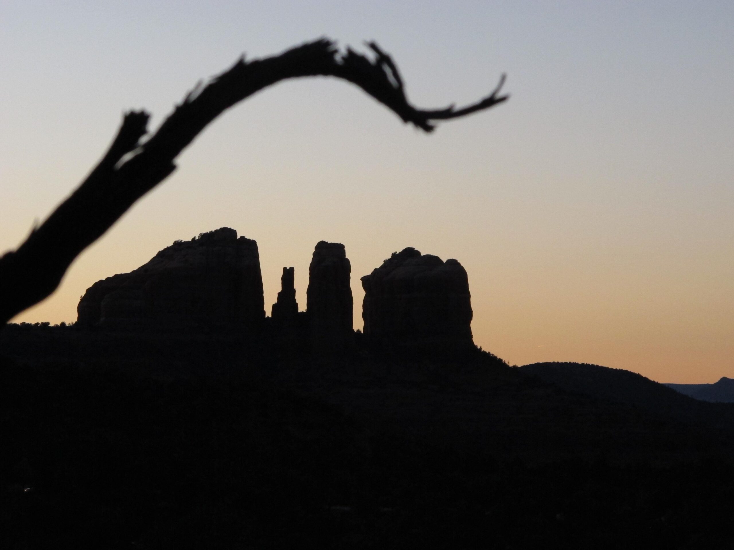 Silhouette of rugged rock formations against a twilight sky, with a branch in the foreground. The colors transition from deep blue to warm orange as the sun sets on the horizon. Bell Rock Area Trails mountain bike trail.