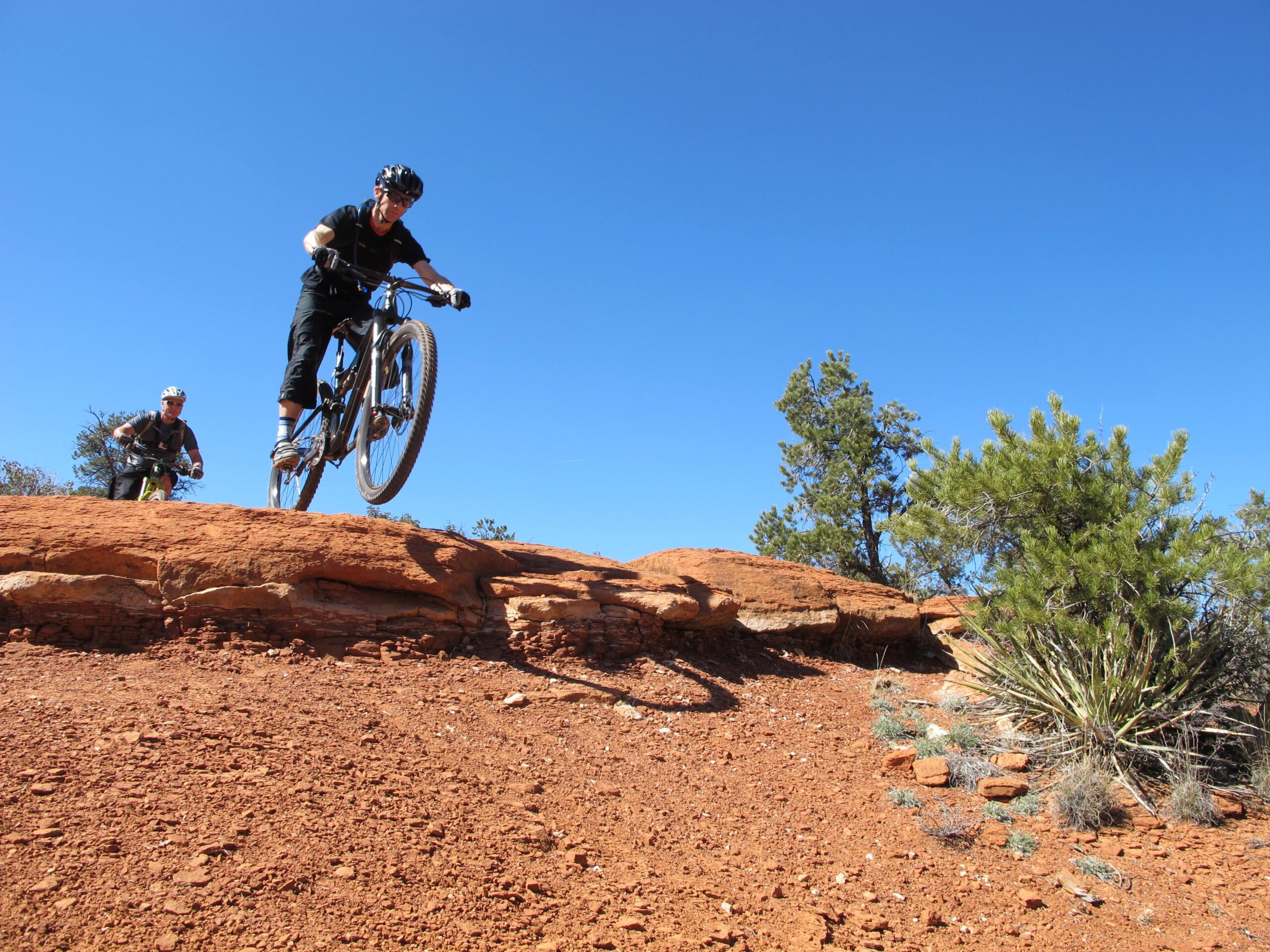 A mountain biker performing a jump over a rocky outcrop, with a second biker riding in the background. The scene features a clear blue sky and desert vegetation, highlighting the rugged terrain. Bell Rock Area Trails mountain bike trail.