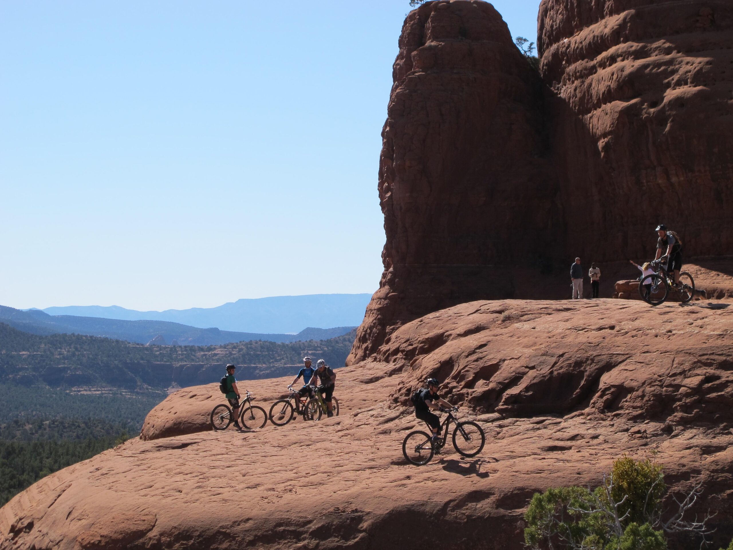 A group of mountain bikers enjoys a ride on a rocky landscape with distant mountains under a clear blue sky. The scene features three cyclists on the left side and two more in the background, while two onlookers observe from a higher rocky area. Bell Rock Area Trails mountain bike trail.