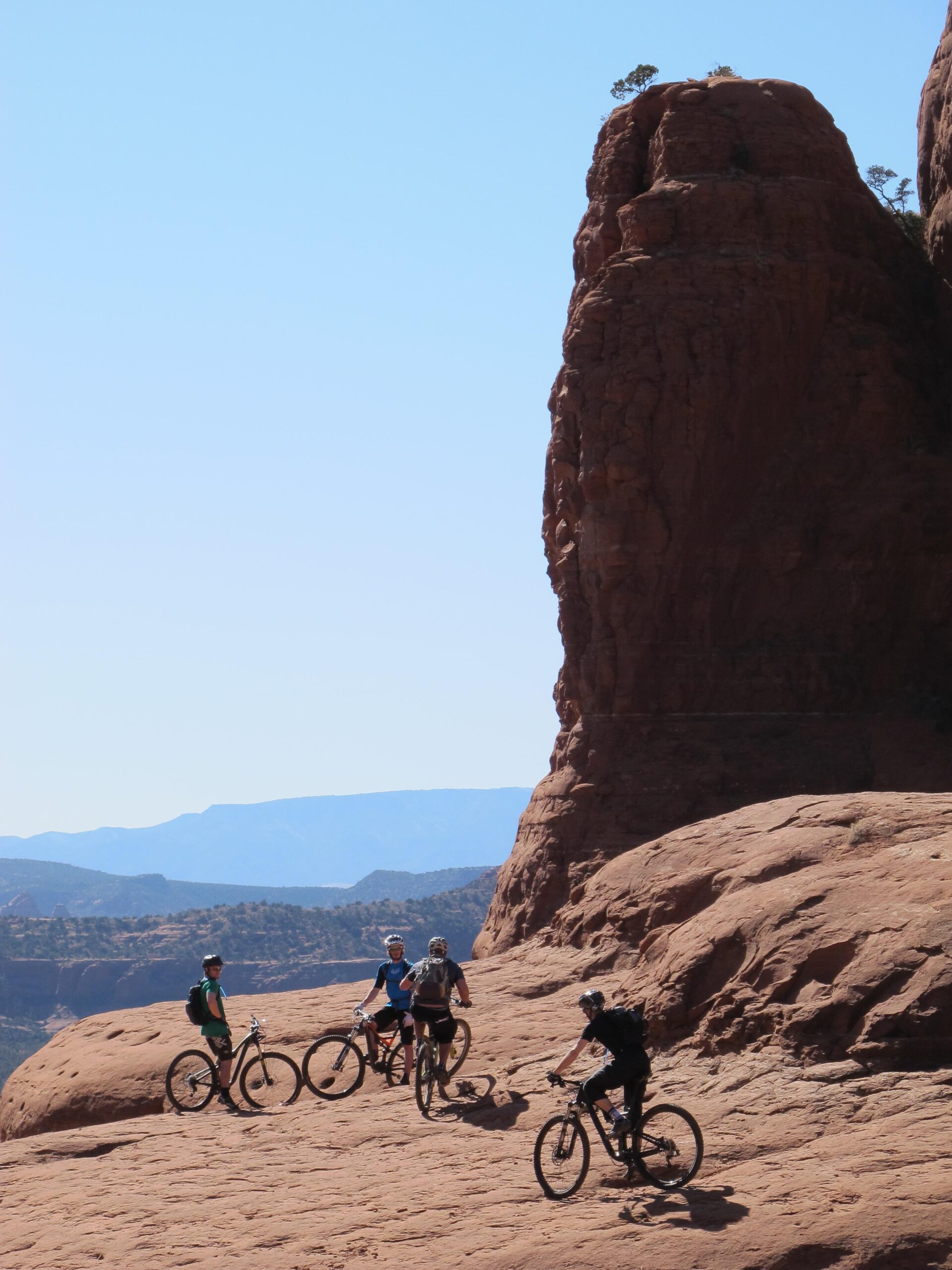 A group of four mountain bikers is riding and standing on rocky terrain, with a large red rock formation in the background. The landscape features layered rocks and distant mountains under a clear blue sky. Bell Rock Area Trails mountain bike trail.