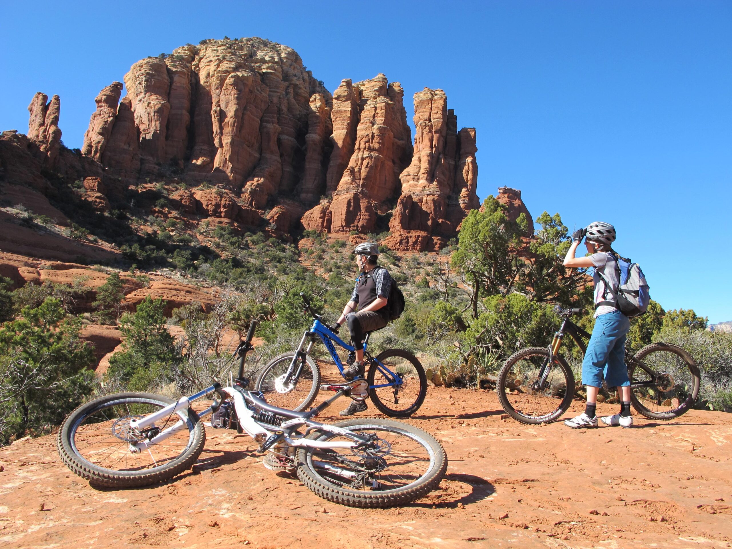 Two mountain bikers stop to take in the view of towering red rock formations under a clear blue sky. One biker stands next to a black mountain bike, while the other adjusts their helmet and gazes into the distance. Two other bikes are lying on the ground nearby, surrounded by sparse vegetation on the reddish terrain. Bell Rock Area Trails mountain bike trail.