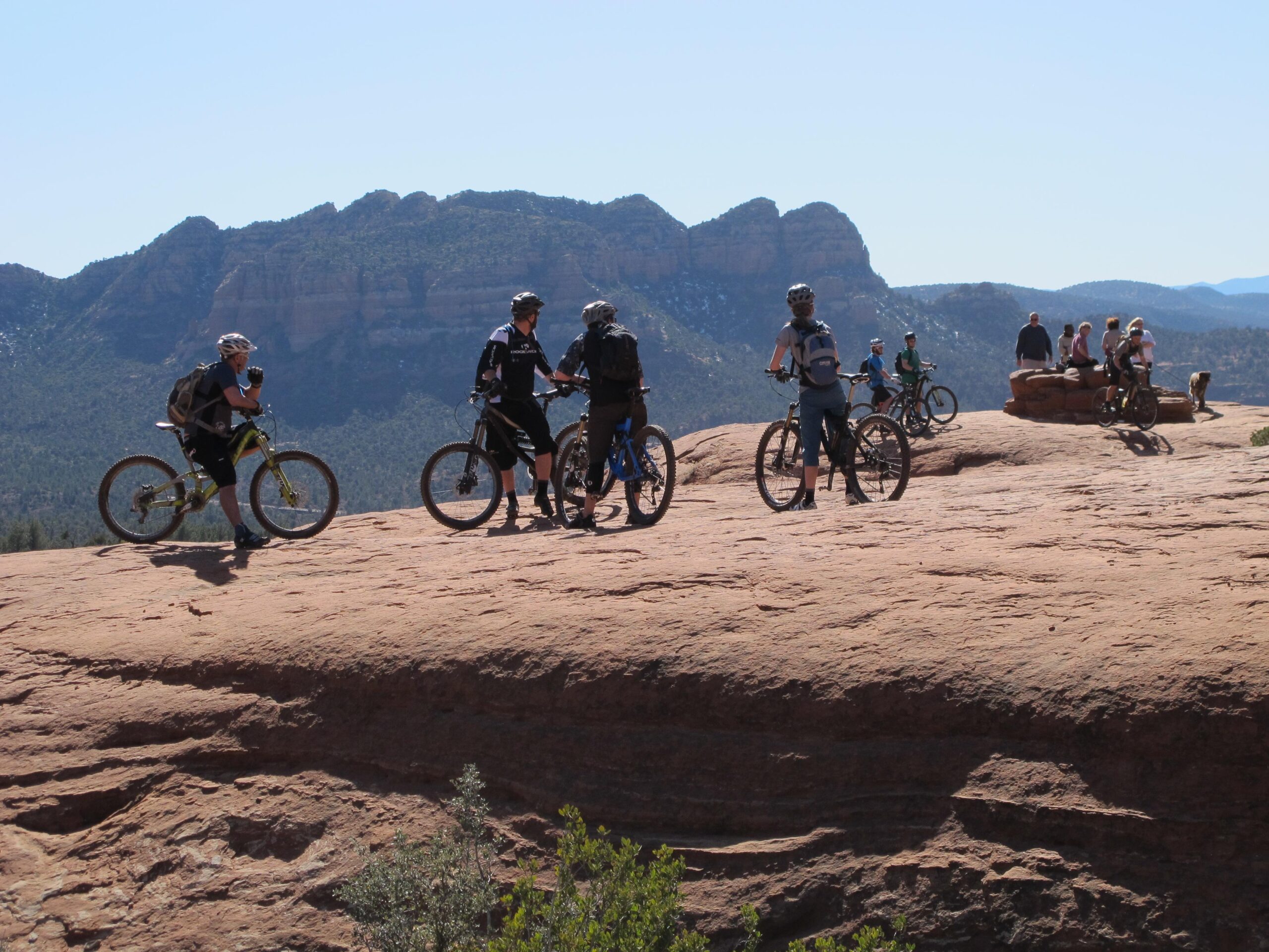 A group of mountain bikers pause on a rocky outcrop, enjoying the scenic view of distant mountains under a clear blue sky. Some riders are standing with their bikes, while others are seated on a rock formation, surrounded by lush greenery. Bell Rock Area Trails mountain bike trail.