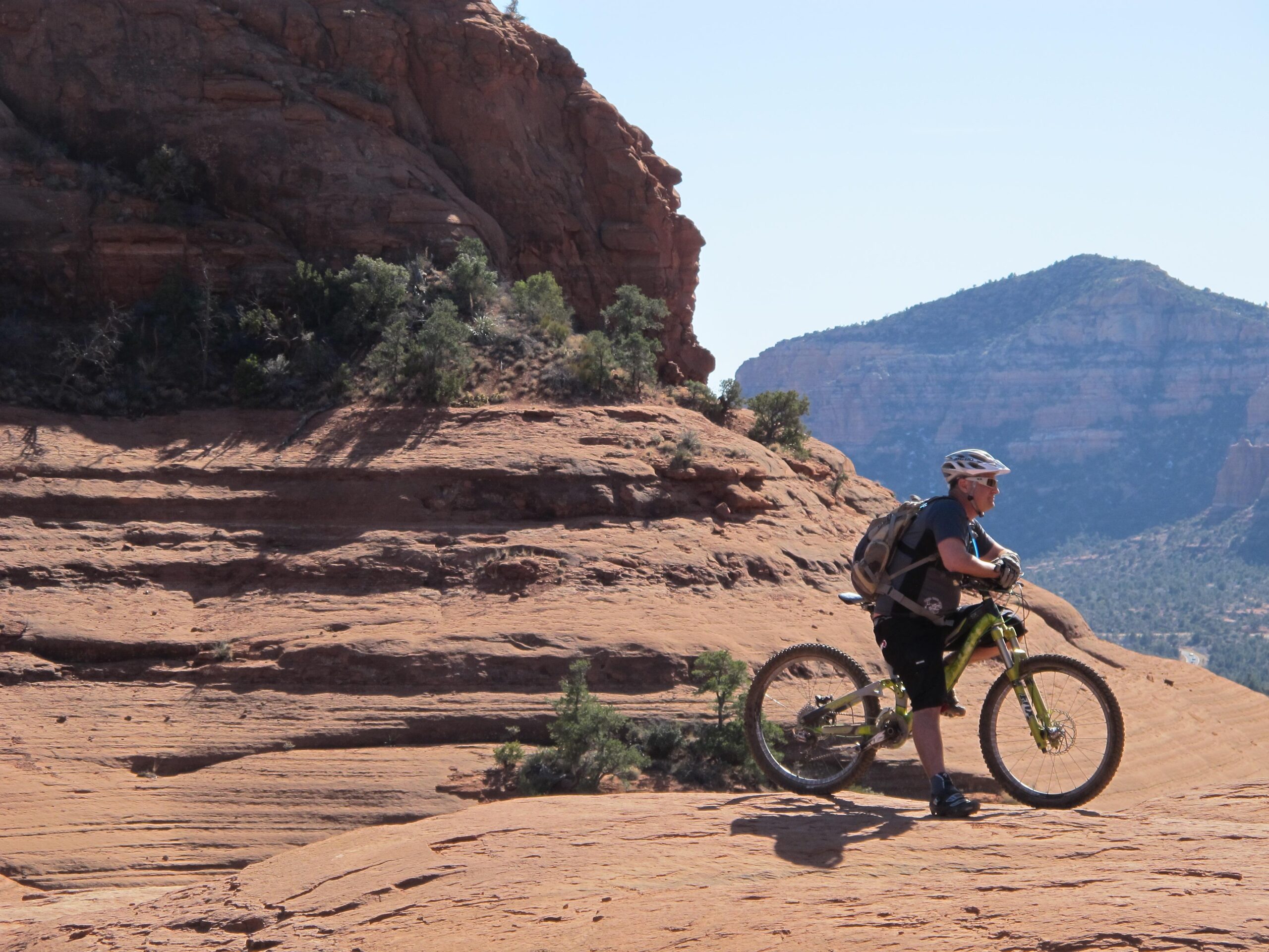 A mountain biker stands next to their bike on a rocky terrain, surrounded by steep, rugged hills and a clear blue sky in the background. The scene reflects a sunny day in a natural outdoor setting, showcasing the beauty of the landscape. Bell Rock Area Trails mountain bike trail.