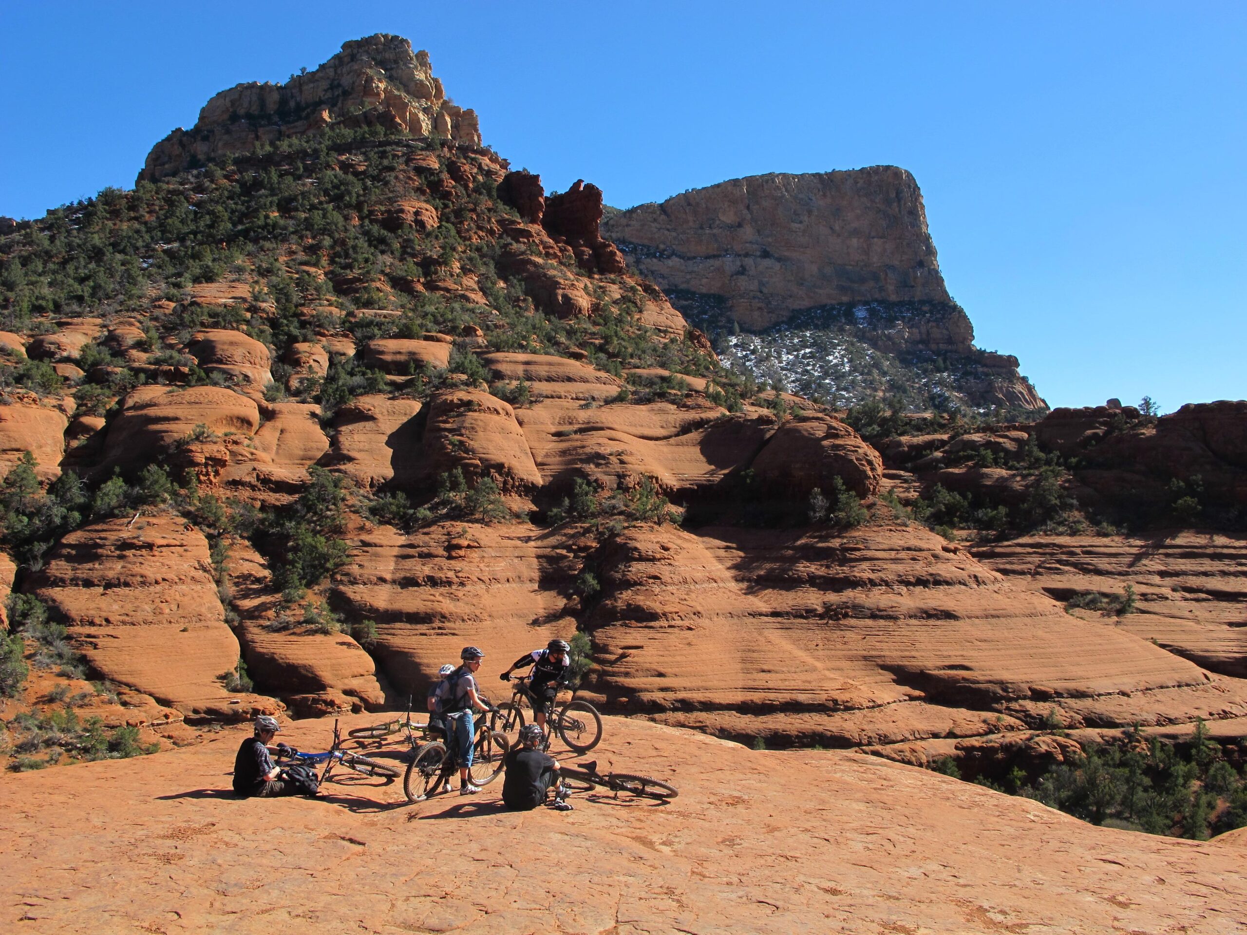 A group of mountain bikers resting on a rocky, red sandstone landscape, with a prominent rock formation and blue sky in the background. Bell Rock Area Trails mountain bike trail.
