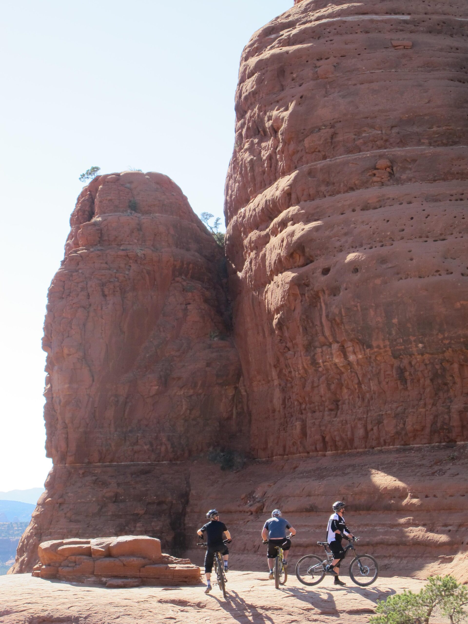 Three mountain bikers stand on rocky terrain, facing a large, towering red rock formation. The sky is clear, and the sun casts shadows across the landscape. The bikers are dressed in casual athletic wear and helmets, with their bicycles beside them. Surrounding vegetation is sparse, emphasizing the rugged beauty of the desert environment. Bell Rock Area Trails mountain bike trail.