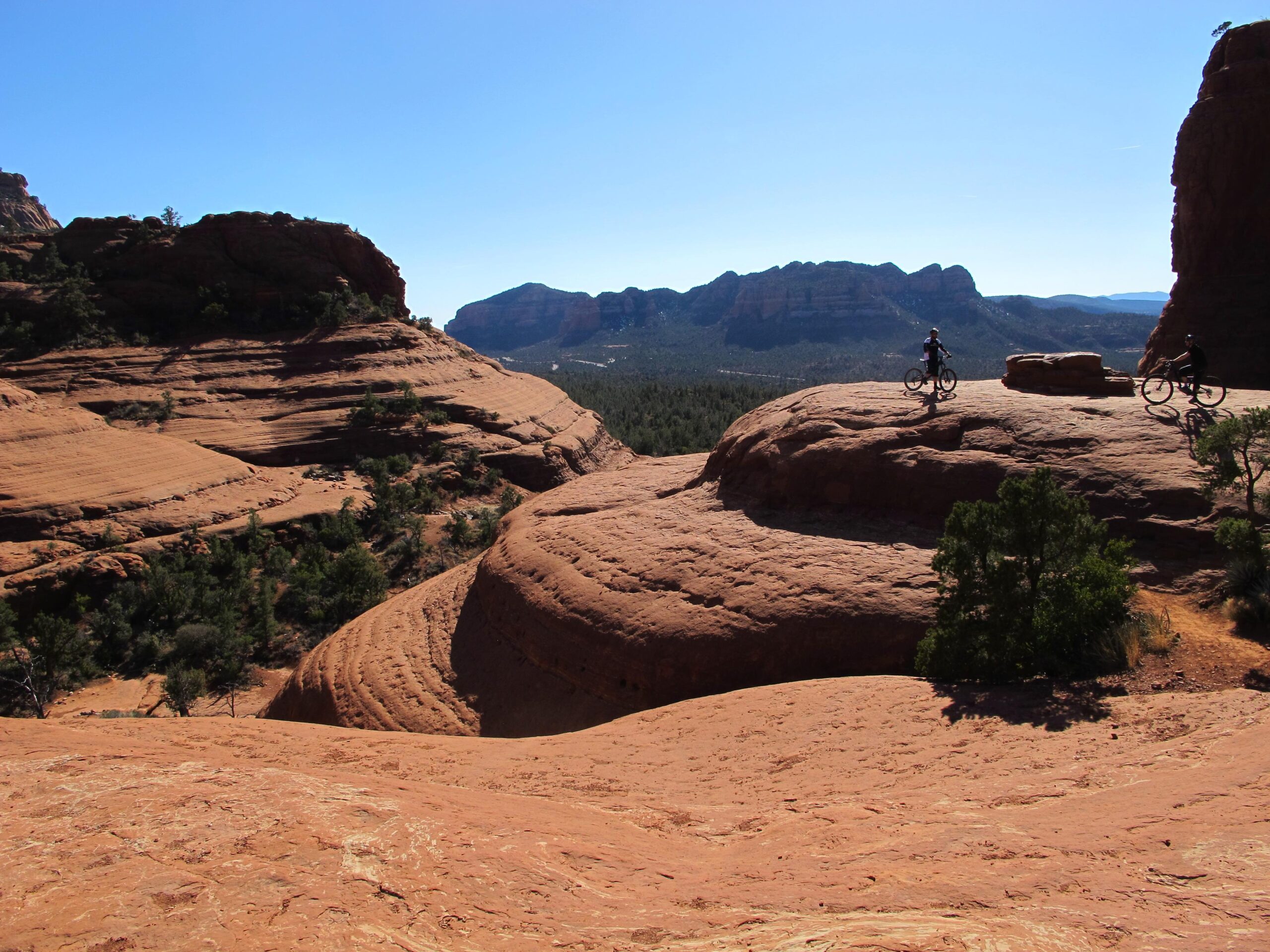 Two mountain bikers on rocky terrain with layered red rock formations and distant mountains under a clear blue sky. Small patches of greenery are visible among the rocks. Bell Rock Area Trails mountain bike trail.