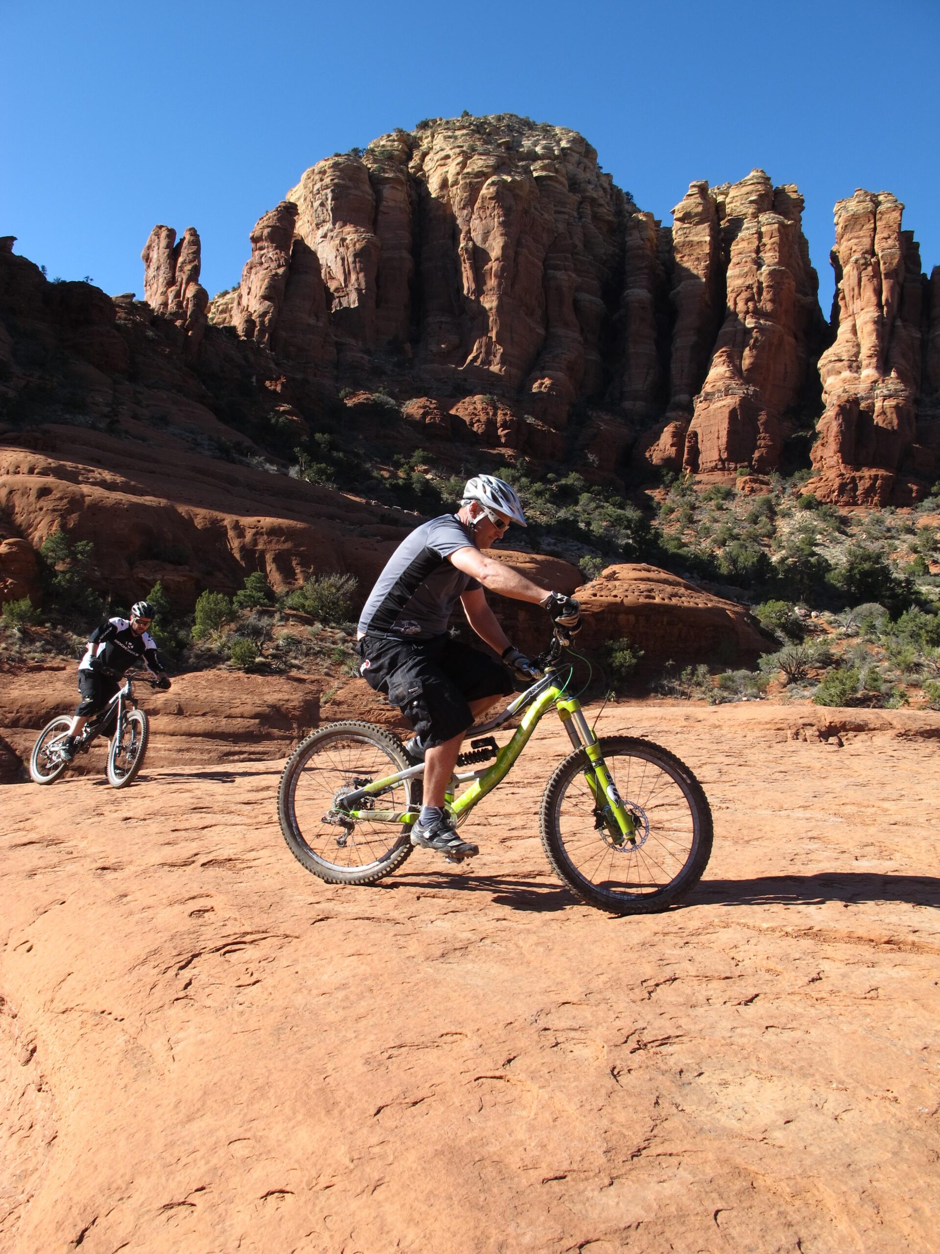 Two mountain bikers navigating a rocky desert trail, with majestic red rock formations and a clear blue sky in the background. One biker is seen closer to the viewer, riding a green bike and wearing a helmet and gray shirt, while the other biker is slightly behind on the trail. Bell Rock Area Trails mountain bike trail.