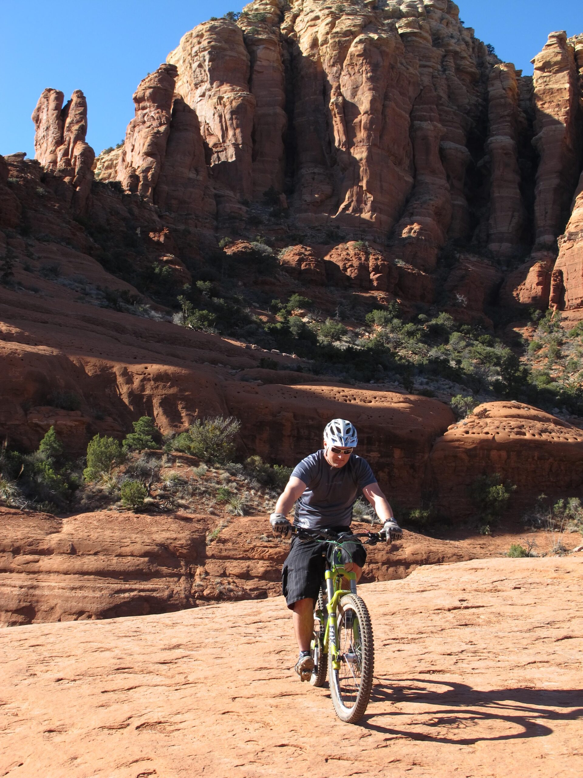 A person riding a green mountain bike on rocky terrain with tall red rock formations and clear blue sky in the background. Bell Rock Area Trails mountain bike trail.