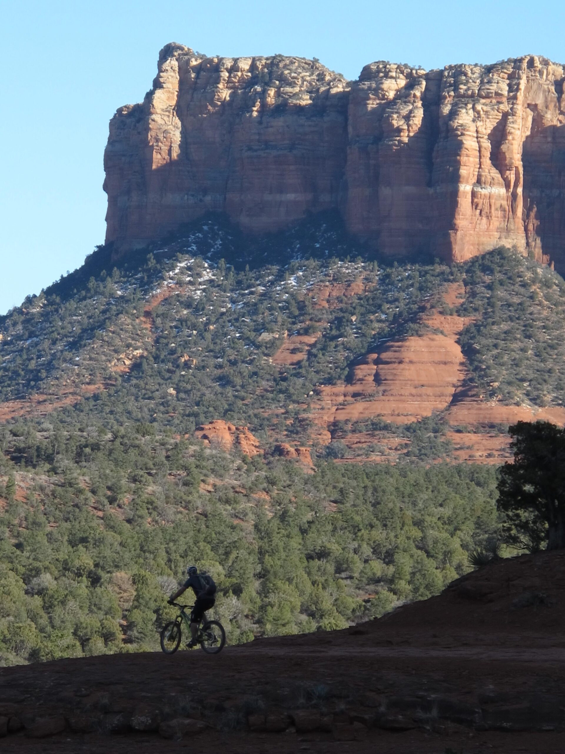 A cyclist riding on a dirt trail in front of a towering red rock formation, surrounded by green pine trees under a clear blue sky. Bell Rock Area Trails mountain bike trail.