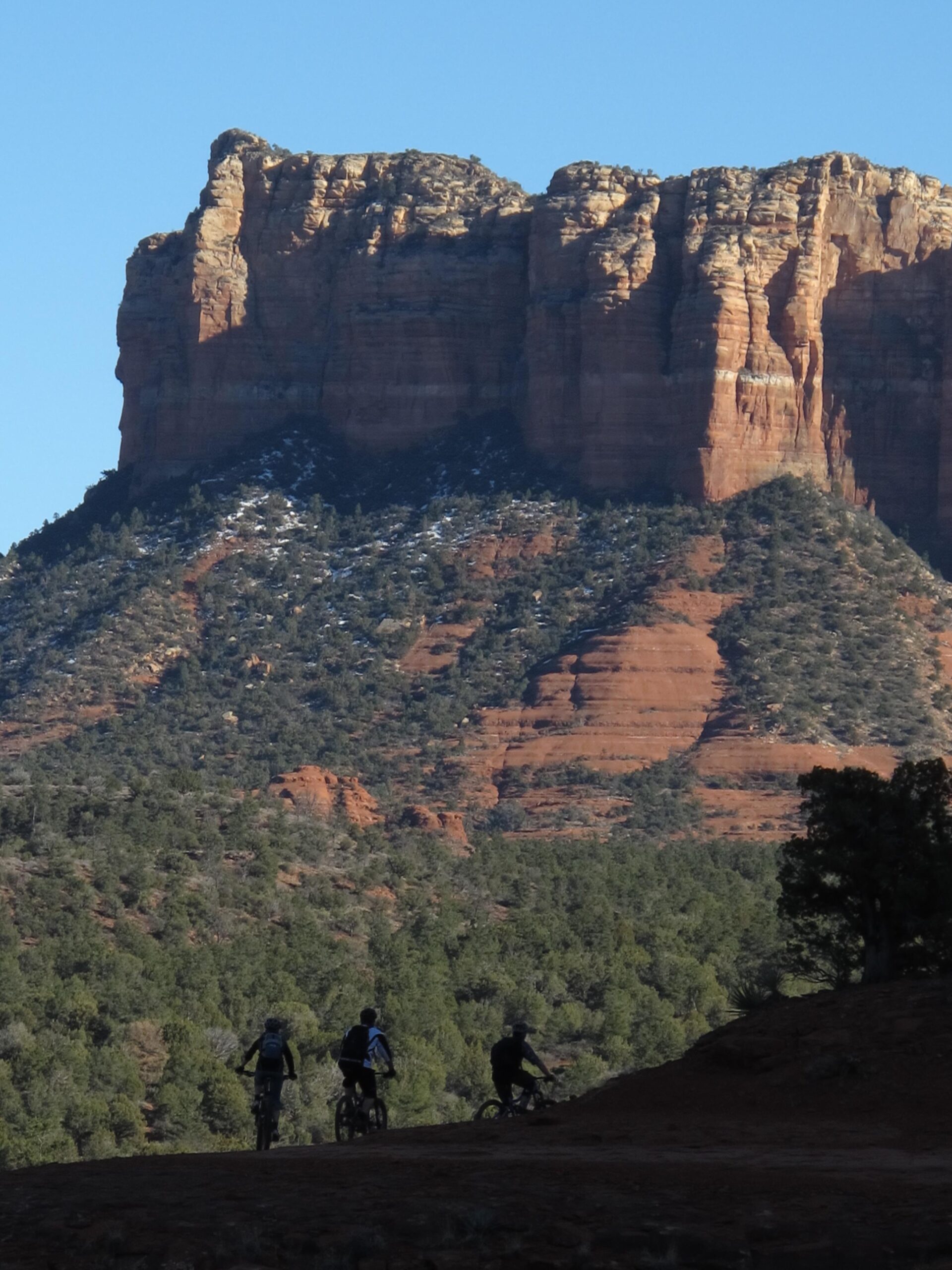 Three mountain bikers ride along a dirt path in front of a large rock formation, surrounded by trees and hills under a clear blue sky. The formation has steep, layered cliffs and a rugged appearance, showcasing the rich red and brown hues typical of the landscape. Bell Rock Area Trails mountain bike trail.
