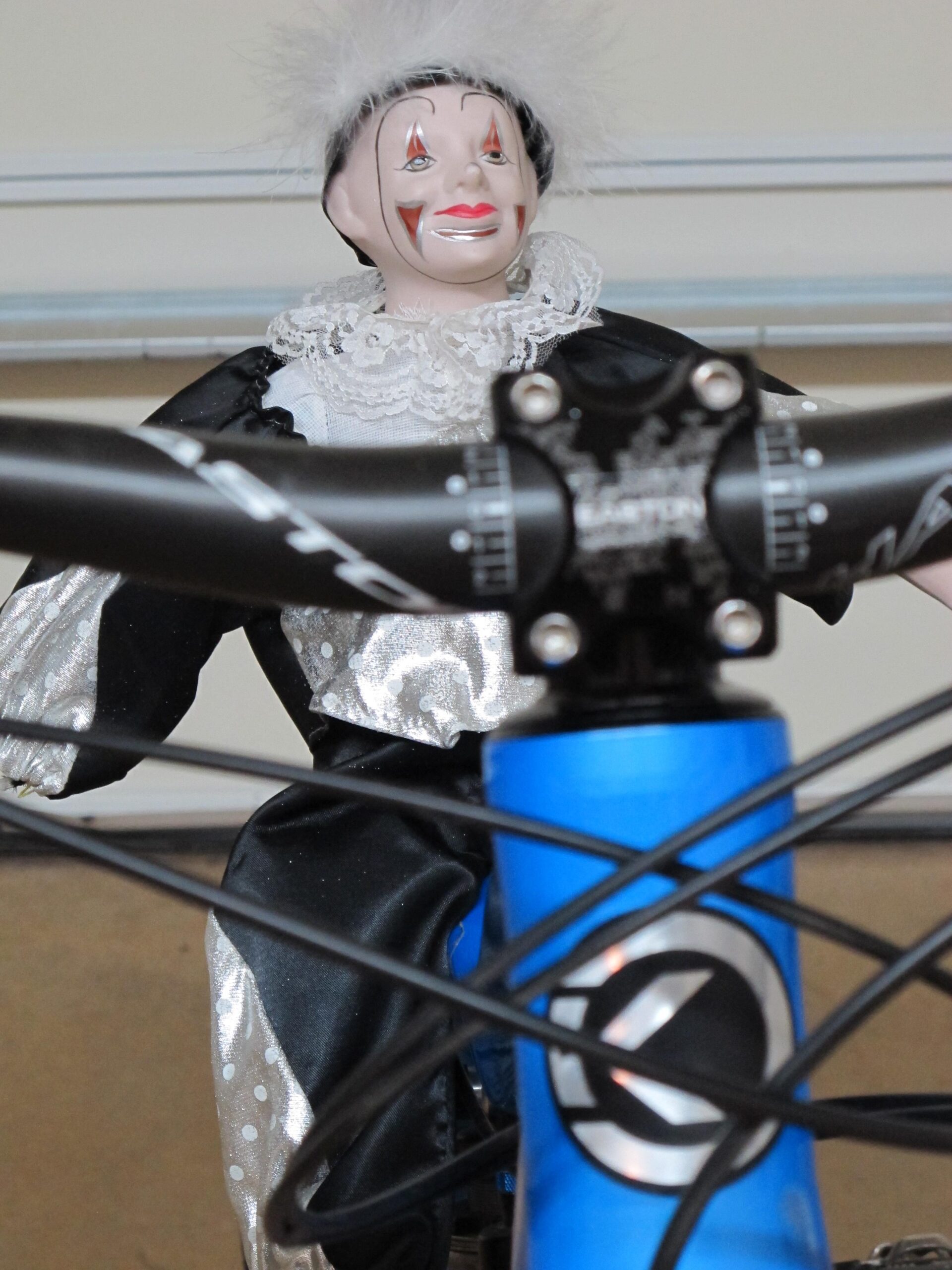 A close-up image of a clown figurine with a white fur hat and a lace collar, sitting on the handlebars of a blue bicycle. The clown has red and black facial paint and is wearing a shiny black and silver costume. Bell Rock Area Trails mountain bike trail.