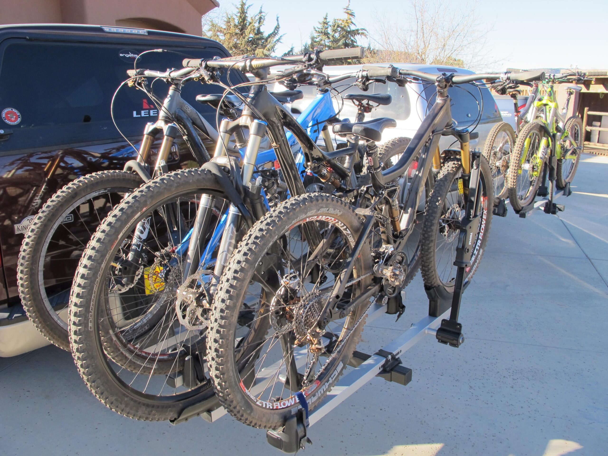 A row of four mountain bikes securely mounted on a bike rack attached to a vehicle, set against a clear sky and a residential backdrop. The bikes vary in color, including black, blue, and green, showcasing their rugged tires and gear components. Bell Rock Area Trails mountain bike trail.