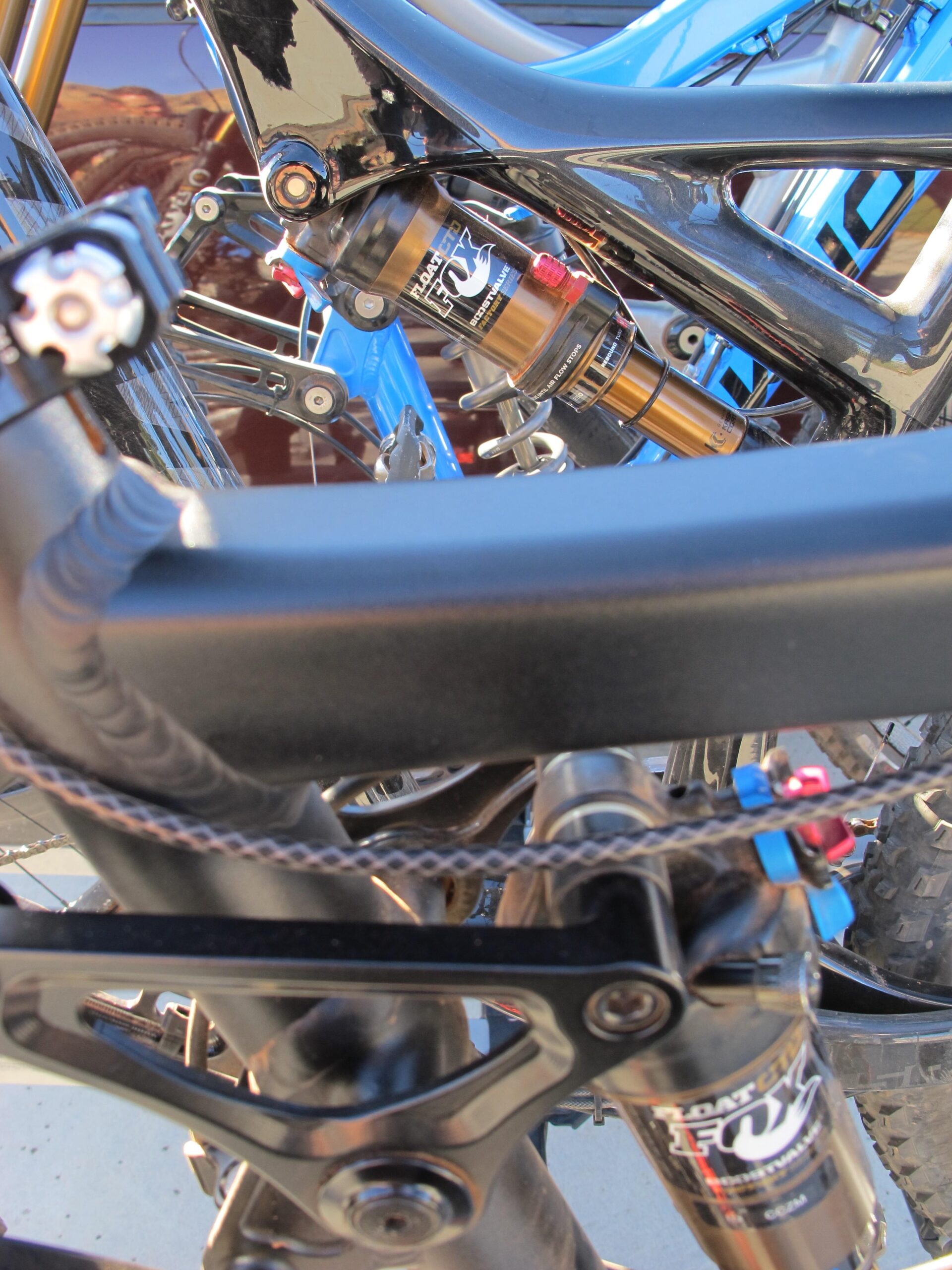 Close-up view of the rear suspension system of a mountain bike, highlighting the black frame, a FOX shock absorber with gold accents, and details of the bike's drivetrain components. The background features a blue frame of another bike, subtly out of focus. Bell Rock Area Trails mountain bike trail.