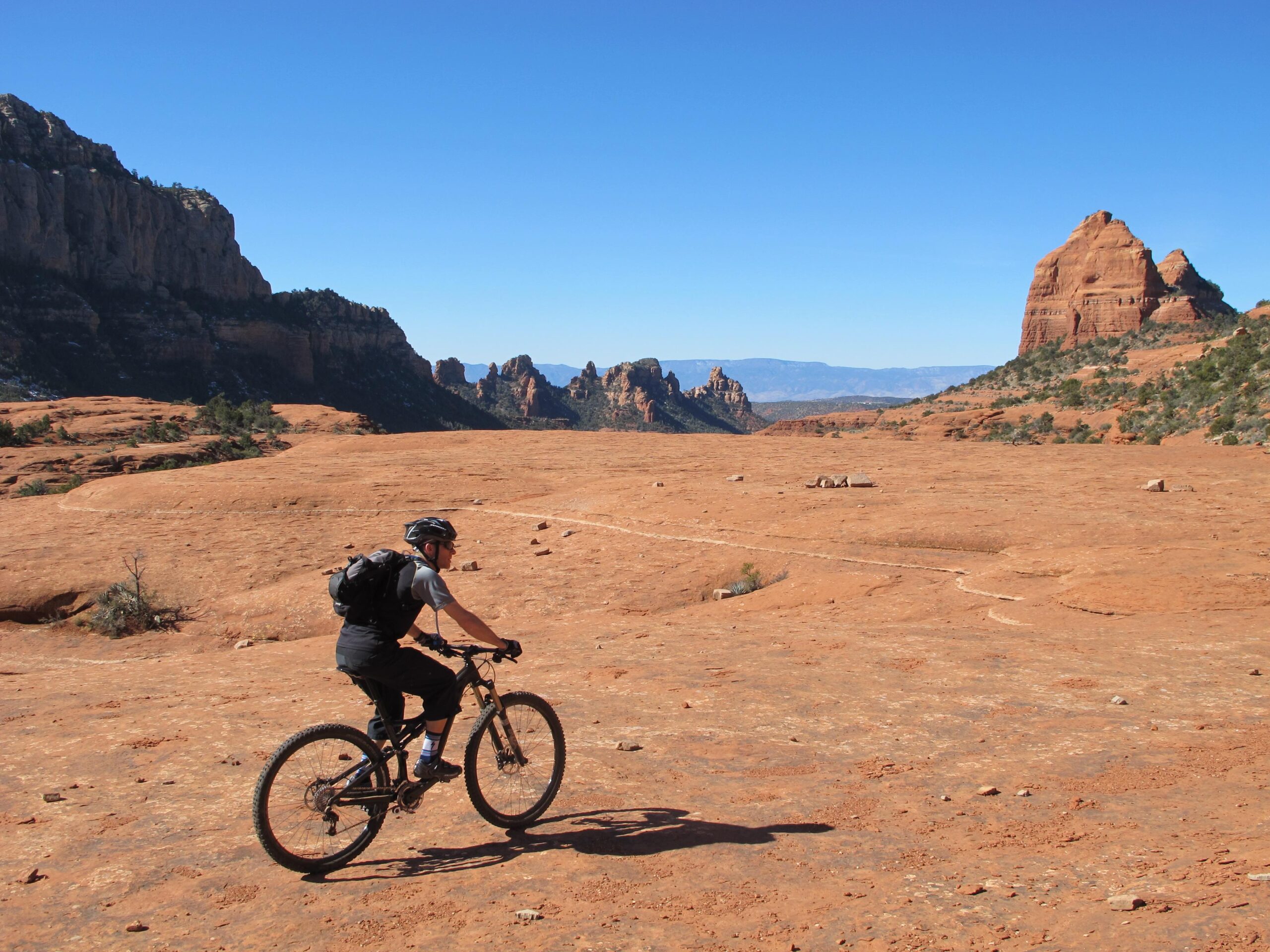 A mountain biker riding on a rocky desert landscape with red sandstone formations and clear blue skies in the background. Bell Rock Area Trails mountain bike trail.
