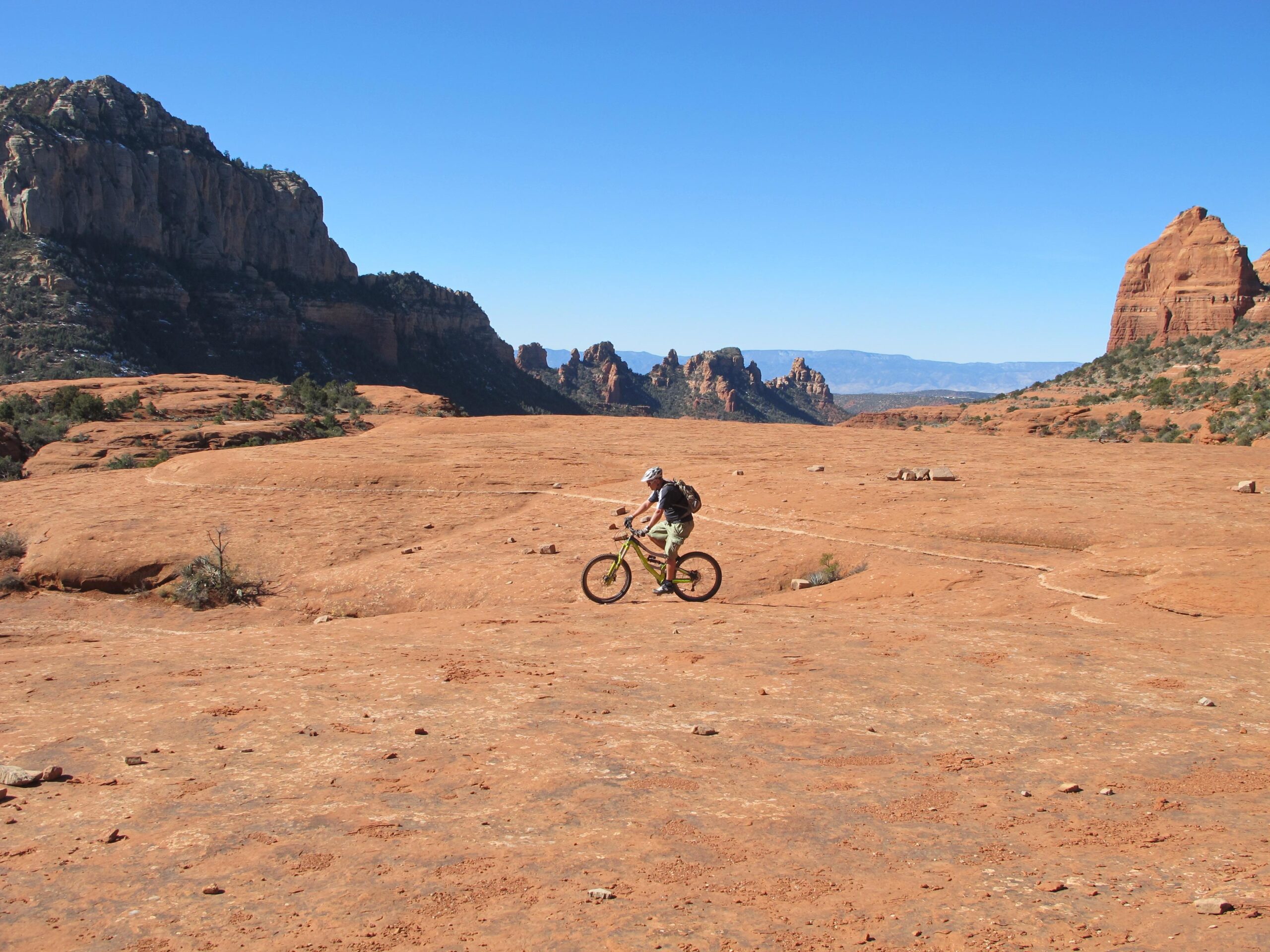 A person riding a mountain bike across a rocky, desert landscape with rugged hills and blue sky in the background. Bell Rock Area Trails mountain bike trail.