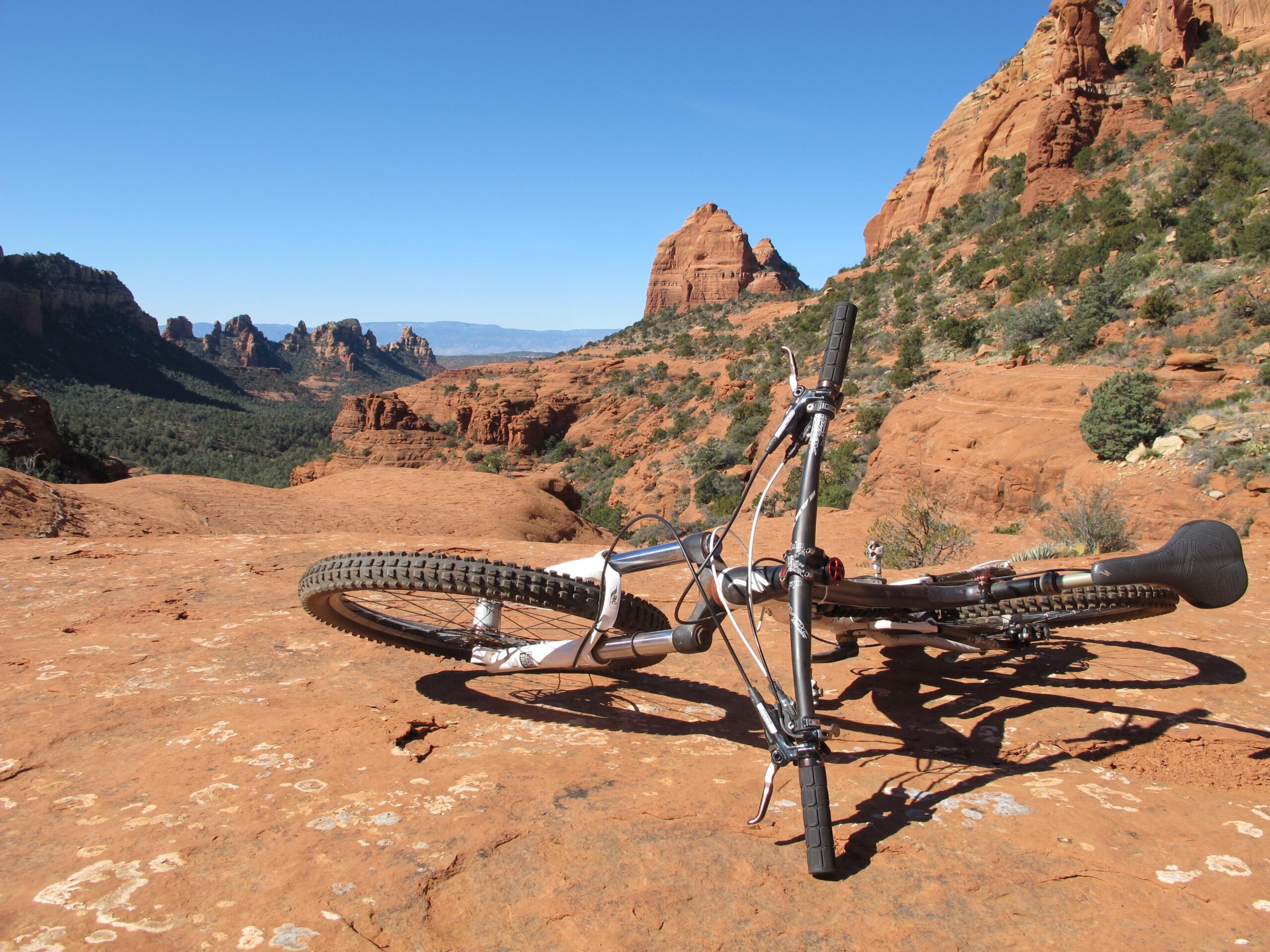 A mountain bike lying on its side on a rocky, reddish terrain, with a panoramic view of rugged hills and blue sky in the background. Bell Rock Area Trails mountain bike trail.