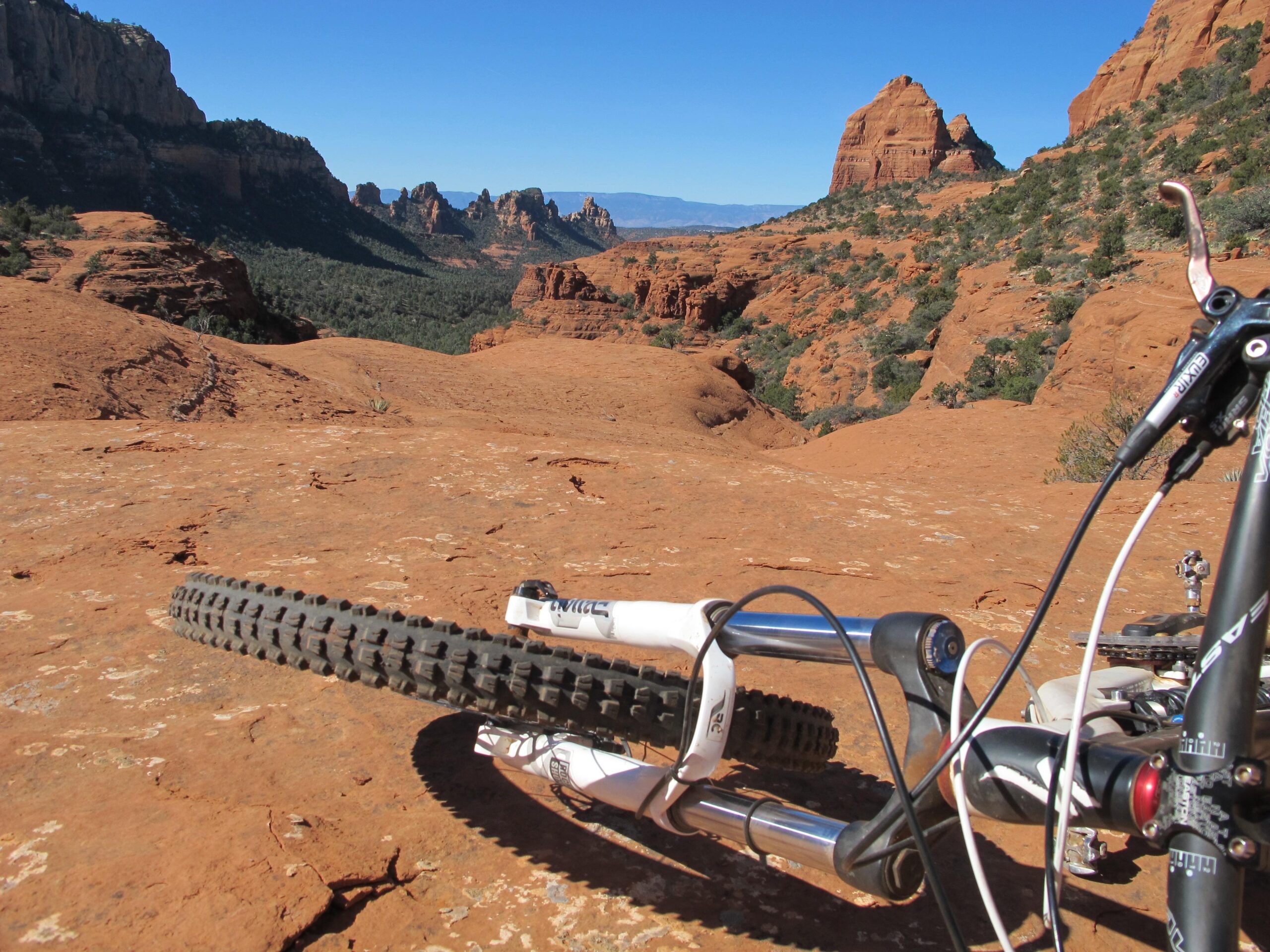 A mountain bike resting on red rock terrain with a scenic backdrop of rugged hills and clear blue skies. The view captures the natural landscape, highlighting the mountain bike's tire and frame in the foreground. Bell Rock Area Trails mountain bike trail.