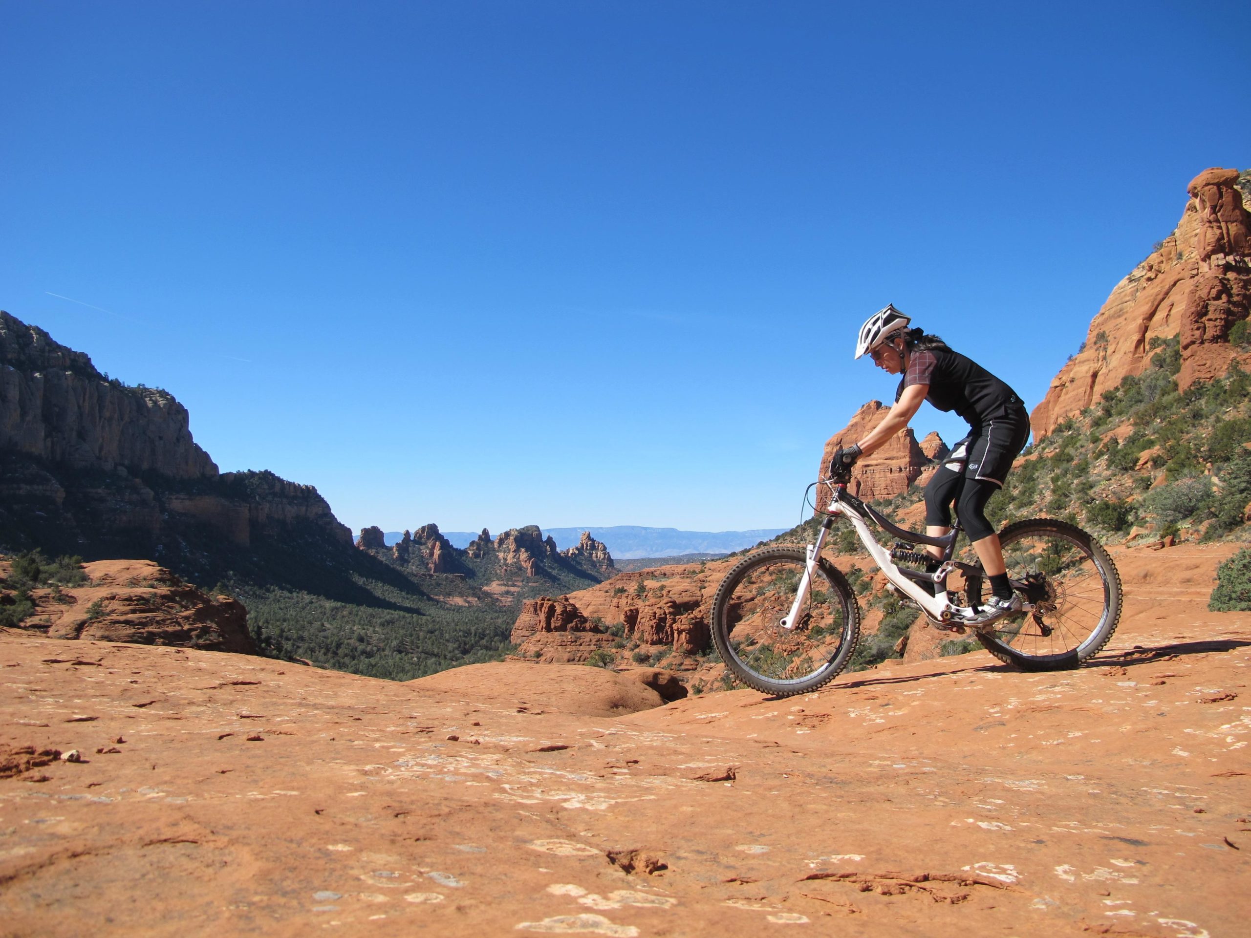 A mountain biker navigates rocky terrain with a clear blue sky in the background, showcasing the dramatic landscape of red rock formations and distant mountains. The cyclist is wearing a helmet and athletic gear, emphasizing an adventurous outdoor activity. Bell Rock Area Trails mountain bike trail.