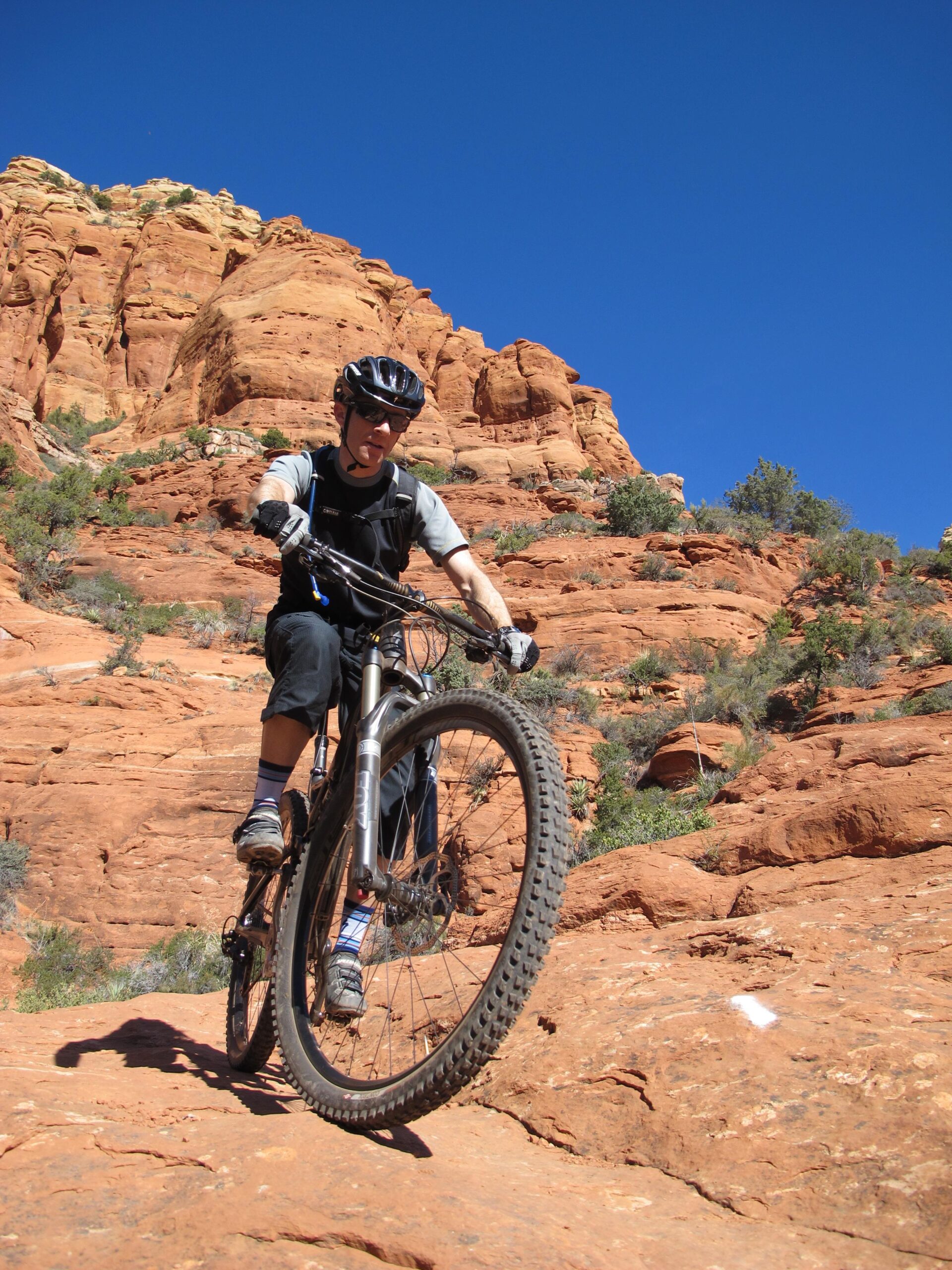A mountain biker navigating rocky terrain under a clear blue sky, with red rock formations in the background. The cyclist is wearing a helmet, sunglasses, and biking gear, concentrating on the challenging trail ahead. Bell Rock Area Trails mountain bike trail.