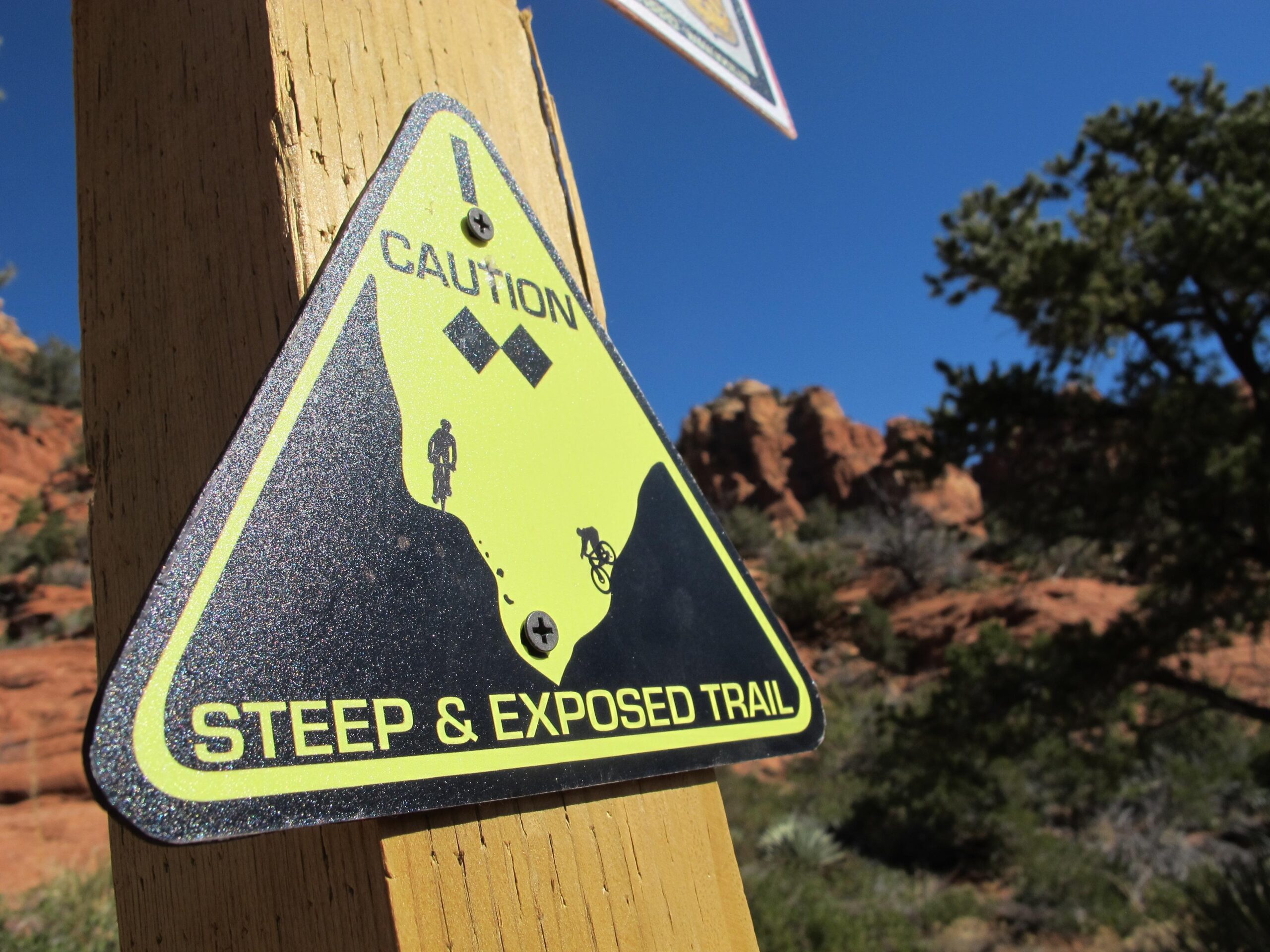 Caution sign on a wooden post indicating a steep and exposed trail, featuring stylized images of a hiker and a cyclist. The background shows a clear blue sky and rocky terrain. Bell Rock Area Trails mountain bike trail.