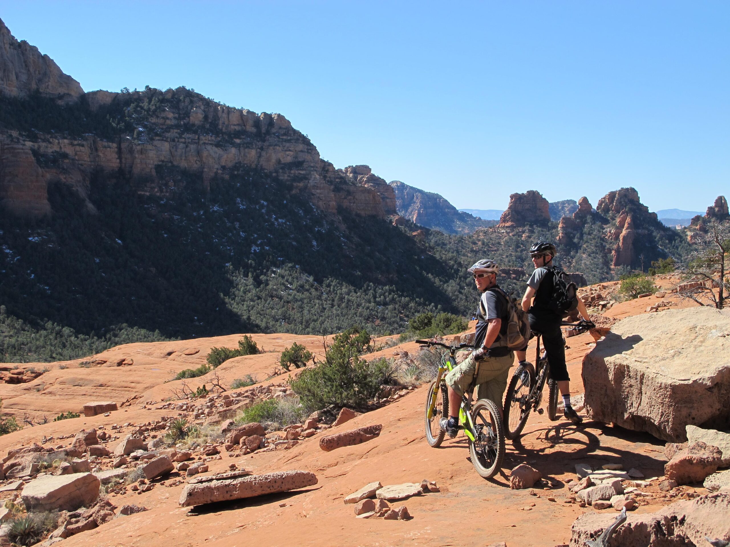 Two mountain bikers pause on a rocky trail, overlooking a scenic view of layered cliffs and greenery in a desert landscape. The sun shines brightly in a clear blue sky. One biker, wearing a helmet and a t-shirt, stands beside his bike, while the other, dressed in shorts and a black shirt, leans on his bike, both enjoying the expansive vista. Bell Rock Area Trails mountain bike trail.
