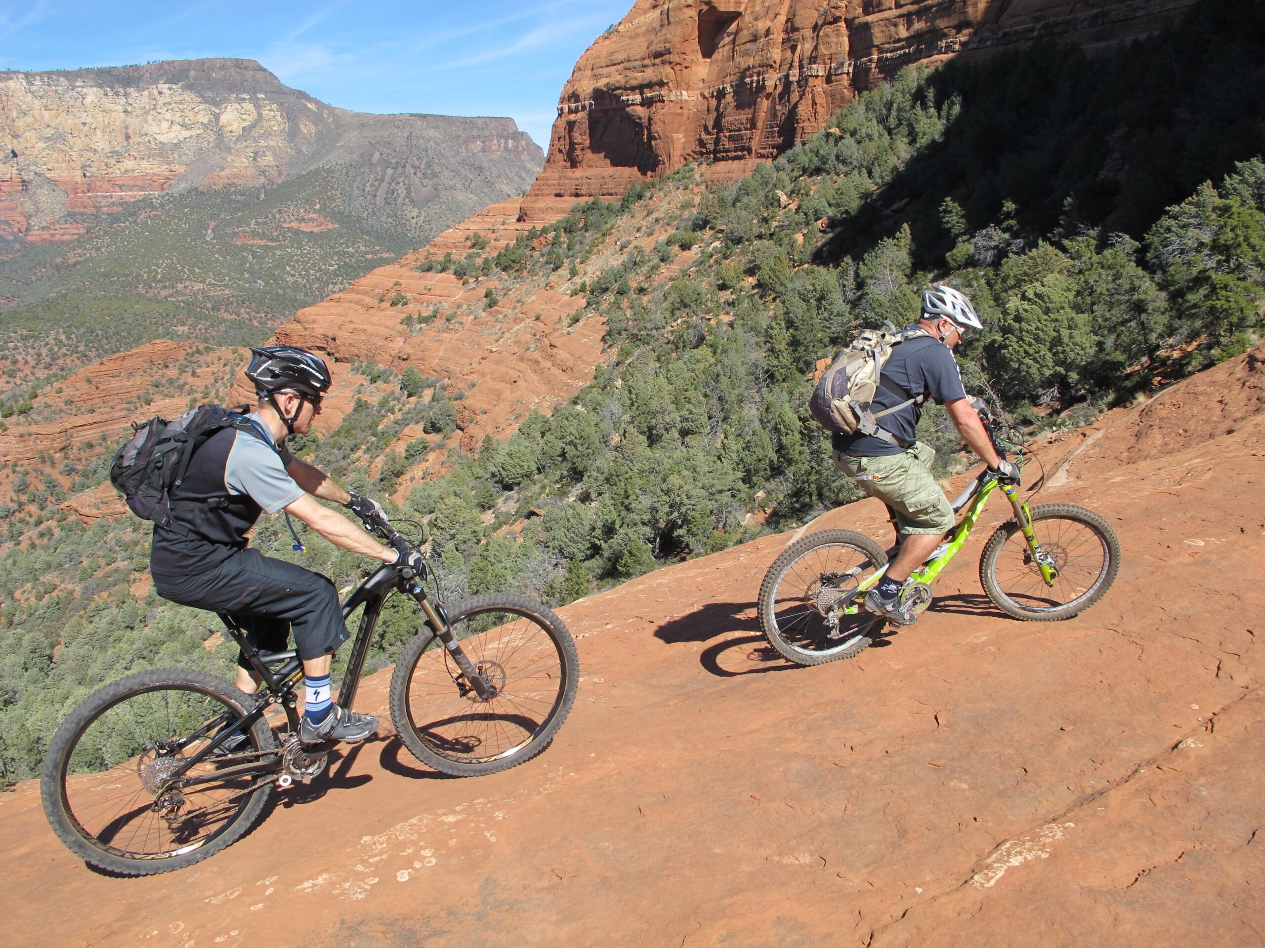 Two mountain bikers navigating a rocky downhill trail, surrounded by rugged red rock formations and green trees. The sky is clear with a few clouds, indicating a sunny day. One rider wears a black helmet and gear, while the other is dressed in a dark shirt and shorts, equipped with a backpack. The landscape shows steep terrain and scenic cliffs in the background. Bell Rock Area Trails mountain bike trail.