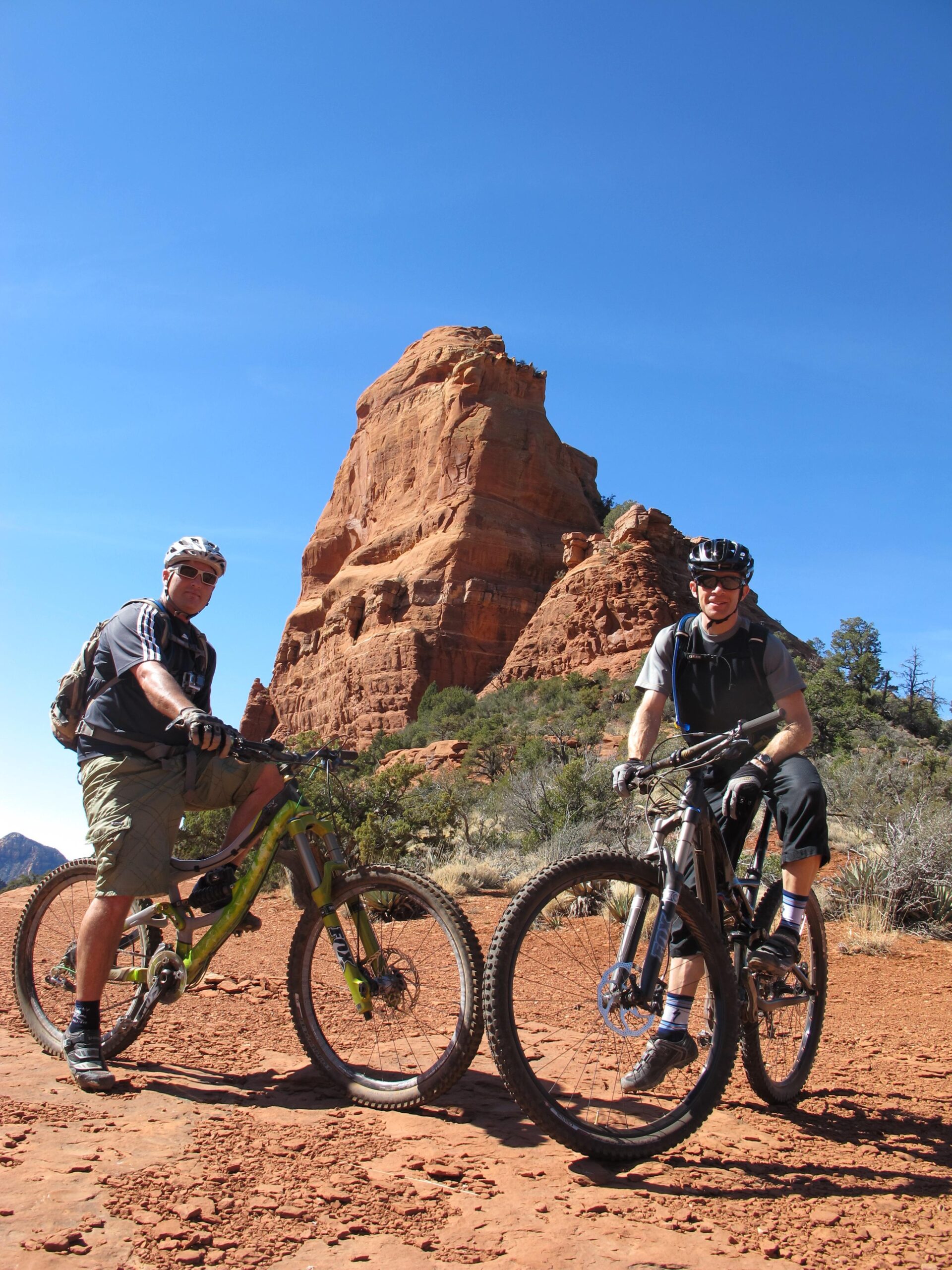 Two mountain bikers pose on a dirt trail against a backdrop of red rock formations under a clear blue sky. One biker, wearing a helmet and sunglasses, stands next to a green bike, while the other biker, dressed in a gray top and black shorts, is seated on a silver bike. Sparse vegetation surrounds the trail, suggesting a sunny outdoor adventure. Bell Rock Area Trails mountain bike trail.