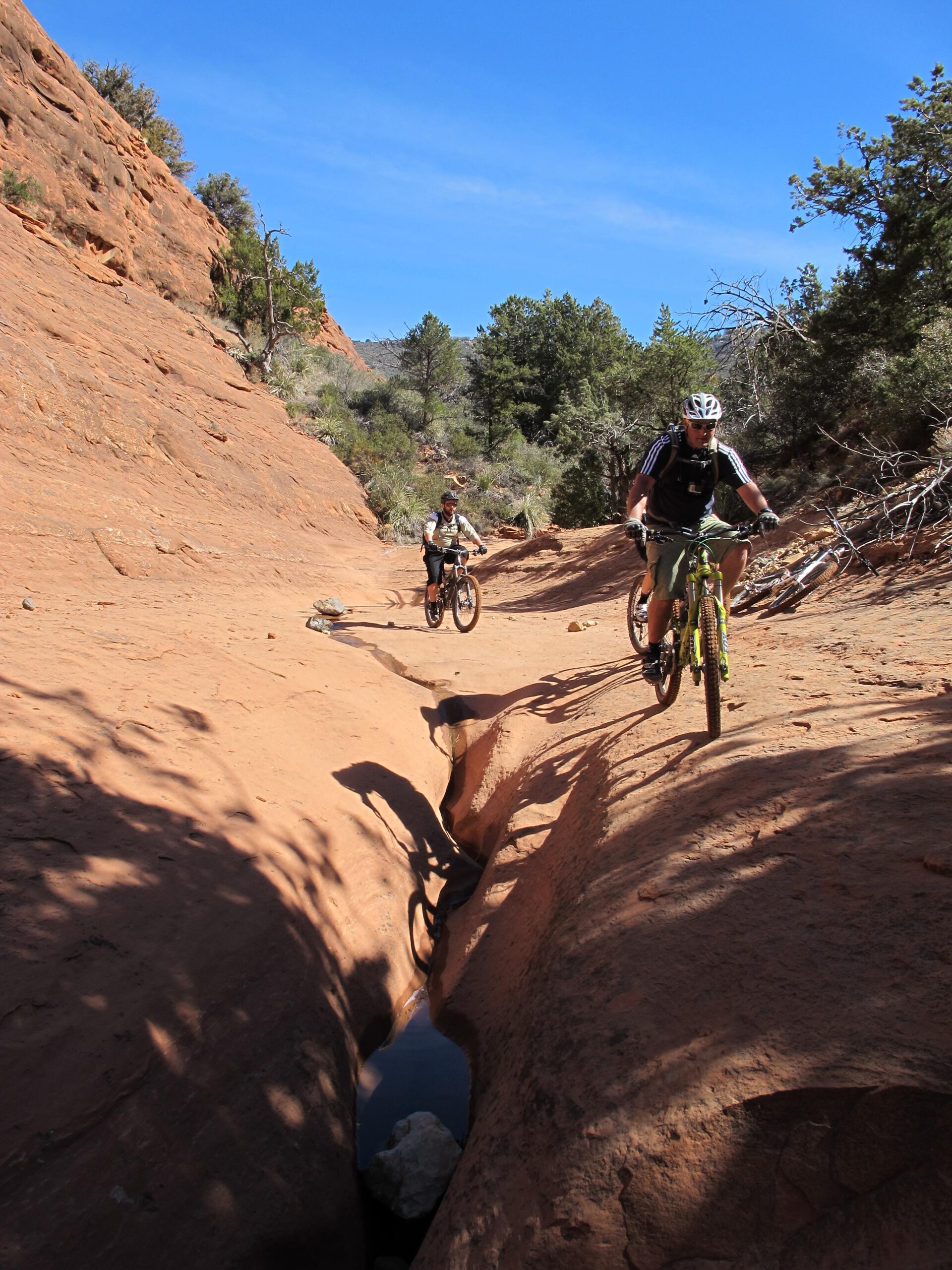 Two mountain bikers navigate a rocky, sandy trail in a canyon-like landscape. The terrain features red rock formations and a narrow channel with water running through it. In the background, trees are visible against a clear blue sky. Bell Rock Area Trails mountain bike trail.