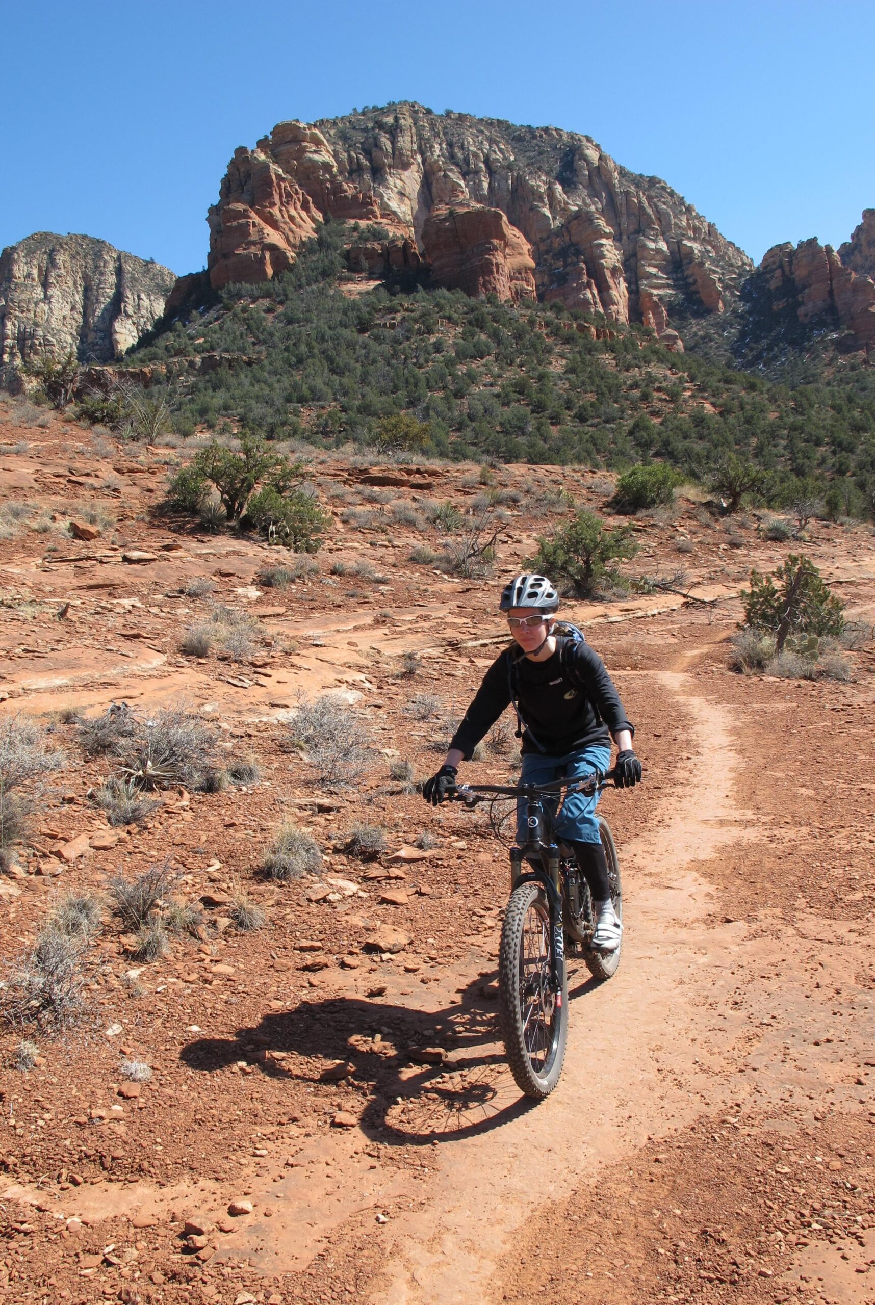 A person riding a mountain bike on a dirt trail in a rocky, desert landscape, with red rock formations and green vegetation in the background under a clear blue sky. Bell Rock Area Trails mountain bike trail.