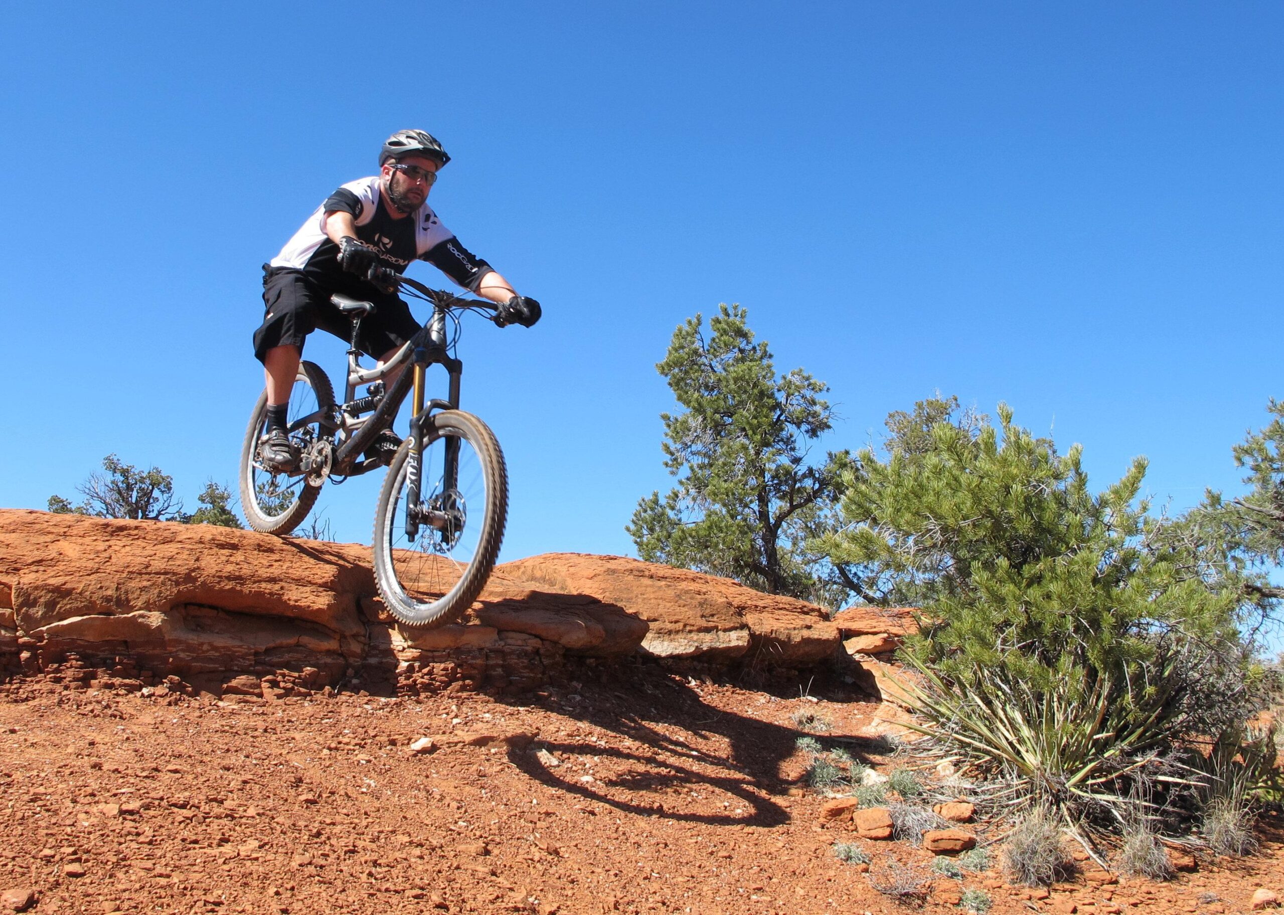 A mountain biker performing a jump over a rocky ledge in a desert landscape, with blue skies and sparse vegetation in the background. Bell Rock Area Trails mountain bike trail.