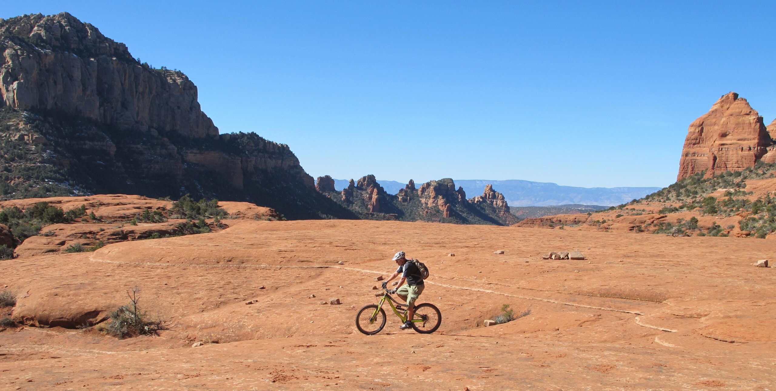 A person riding a mountain bike on a rocky terrain with red sandstone formations and clear blue skies in the background. Bell Rock Area Trails mountain bike trail.