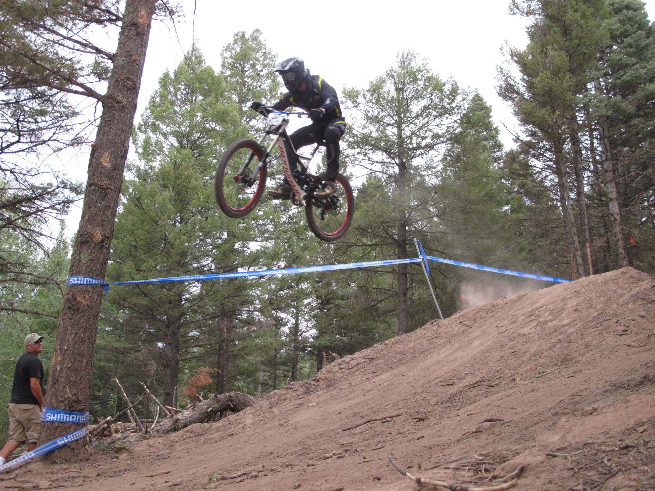 A mountain biker performing a jump off a dirt ramp, airborne with the bike angled under him. A spectator watches from the side, surrounded by trees in a forested area. Blue tape marking the course is visible in the background. Dust is rising from the ramp as the biker lands. Angel Fire Bike Park mountain bike trail.