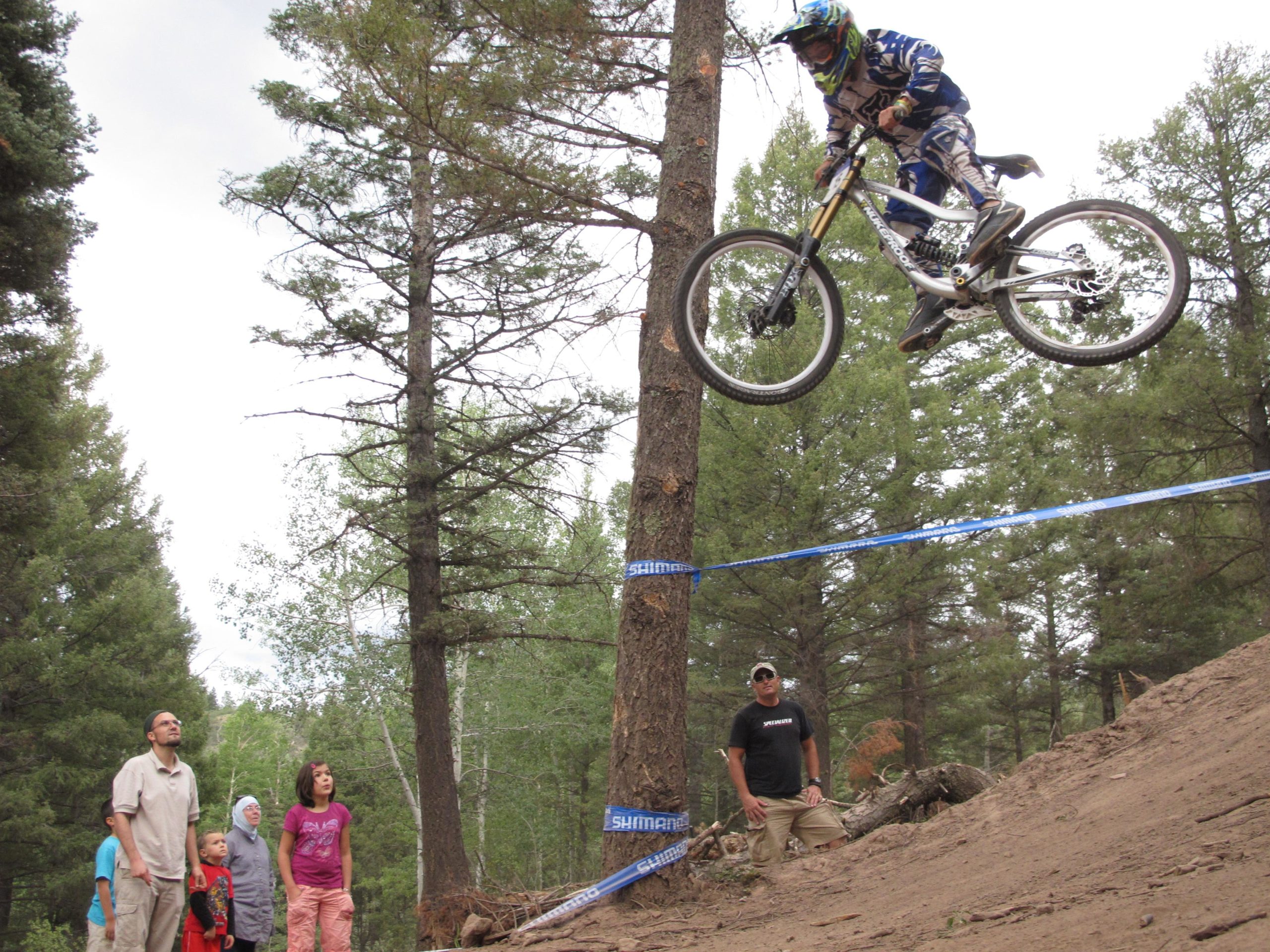 A mountain biker performing a jump off a dirt ramp in a forested area, with spectators watching in the foreground. The biker is wearing a helmet and protective gear, and the scene captures the excitement and energy of the moment. Tall trees and a cloudy sky are visible in the background. Angel Fire Bike Park mountain bike trail.