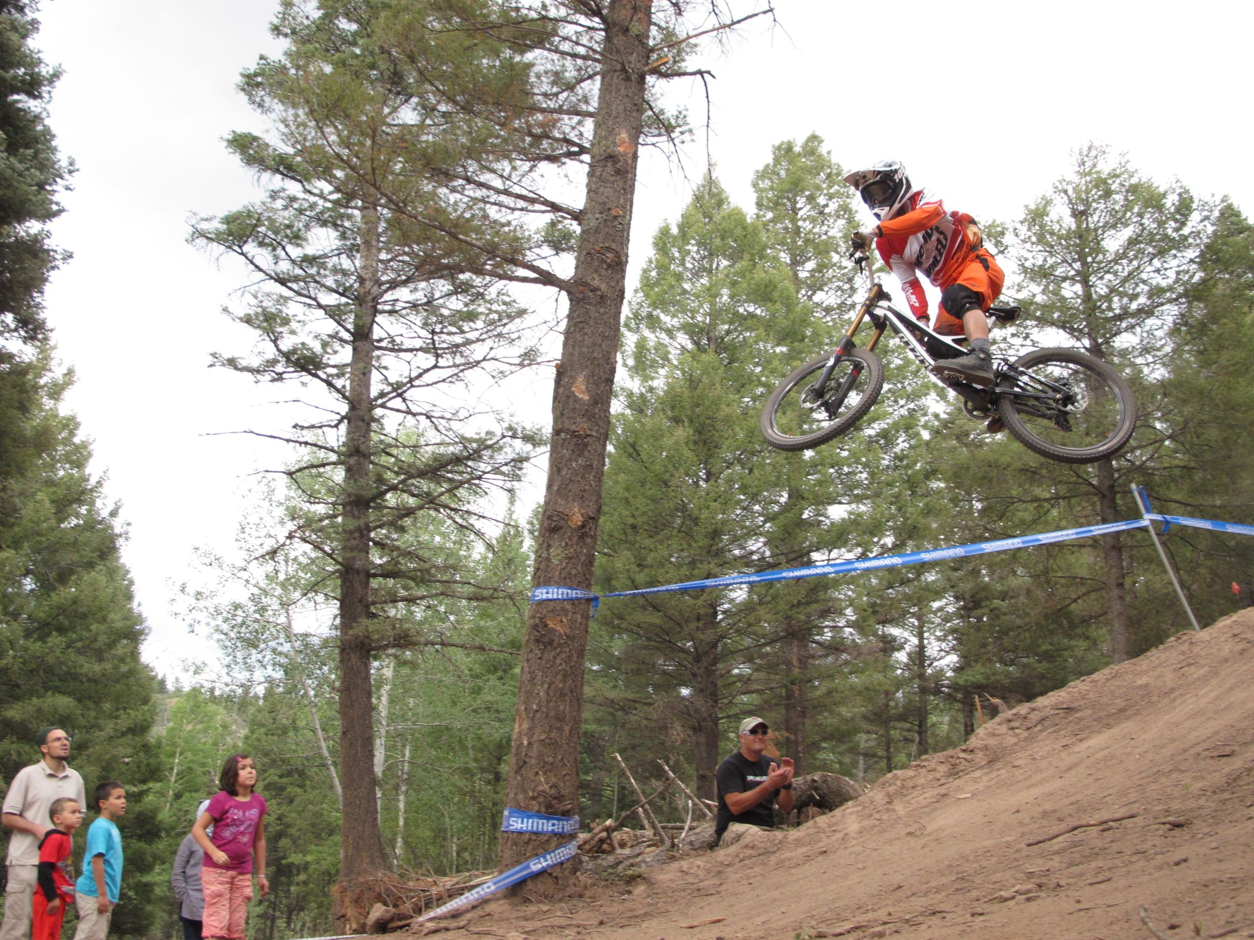 A mountain biker in an orange outfit is performing a jump over a dirt ramp, with trees in the background. Spectators, including children, watch from the side, showing expressions of excitement and attention. Blue tape is visible marking the area. Angel Fire Bike Park mountain bike trail.