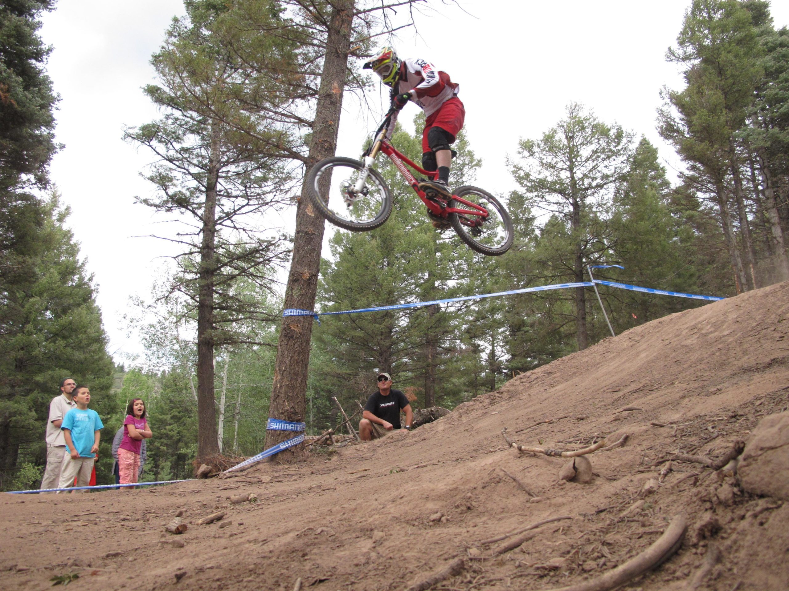 A mountain biker performing a jump over a dirt ramp in a forested area, with spectators watching from the side. Trees surround the scene, and a blue ribbon marks the trail. The rider is wearing a helmet and protective gear, showcasing an action-packed moment in biking. Angel Fire Bike Park mountain bike trail.
