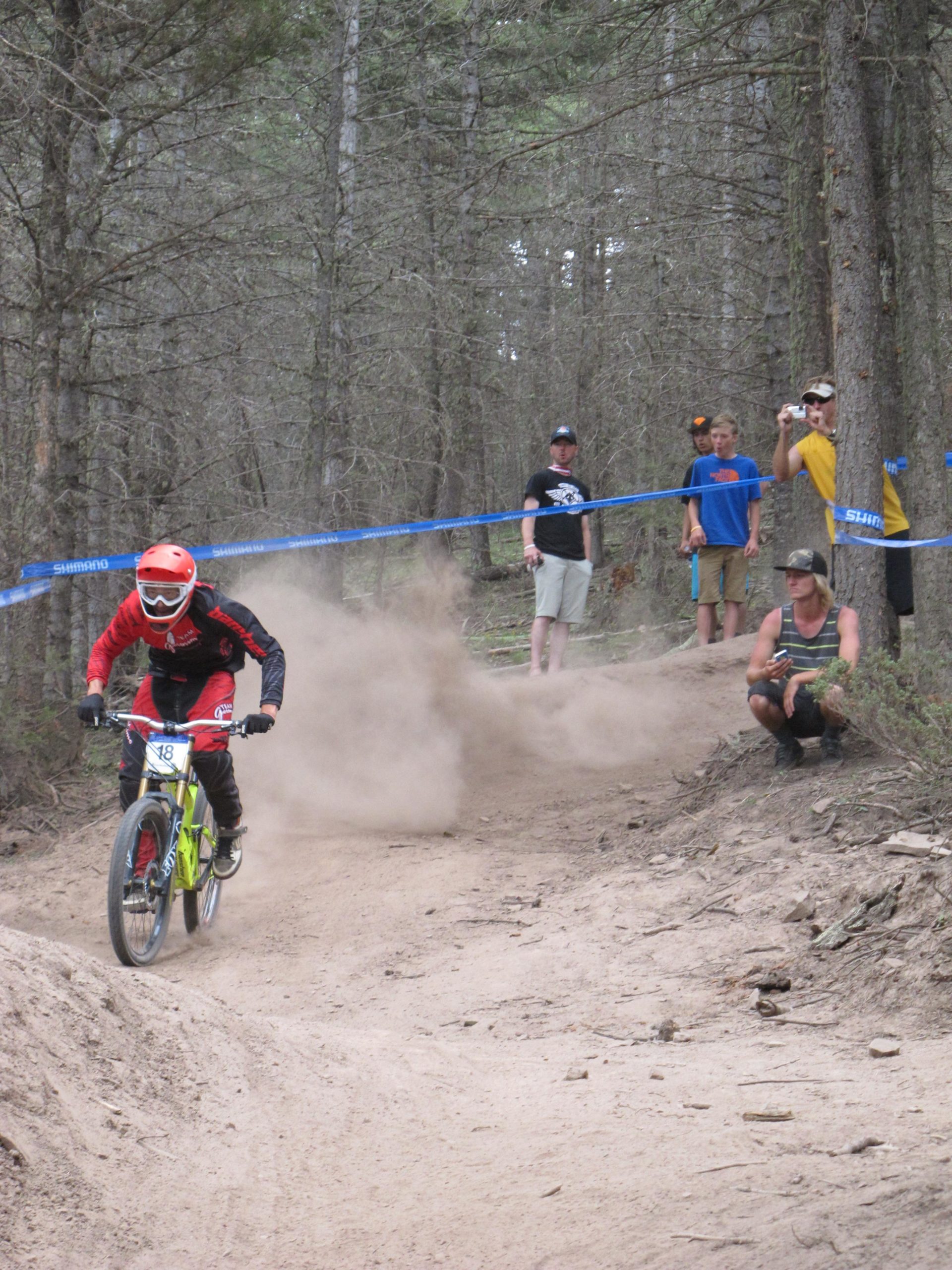 A mountain biker in red and black gear rides along a dirt track, kicking up dust as they navigate a turn. In the background, a small group of spectators watches the race, some holding cameras. The setting is a wooded area with trees lining the trail. Angel Fire Bike Park mountain bike trail.