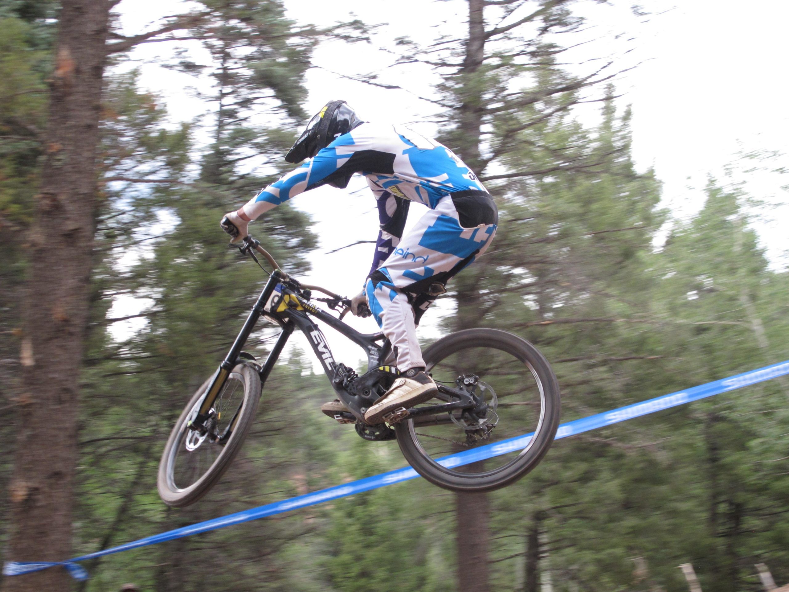 A cyclist in a blue and white racing suit performing a jump on a mountain bike, with trees in the background and a blue banner in the foreground. Angel Fire Bike Park mountain bike trail.