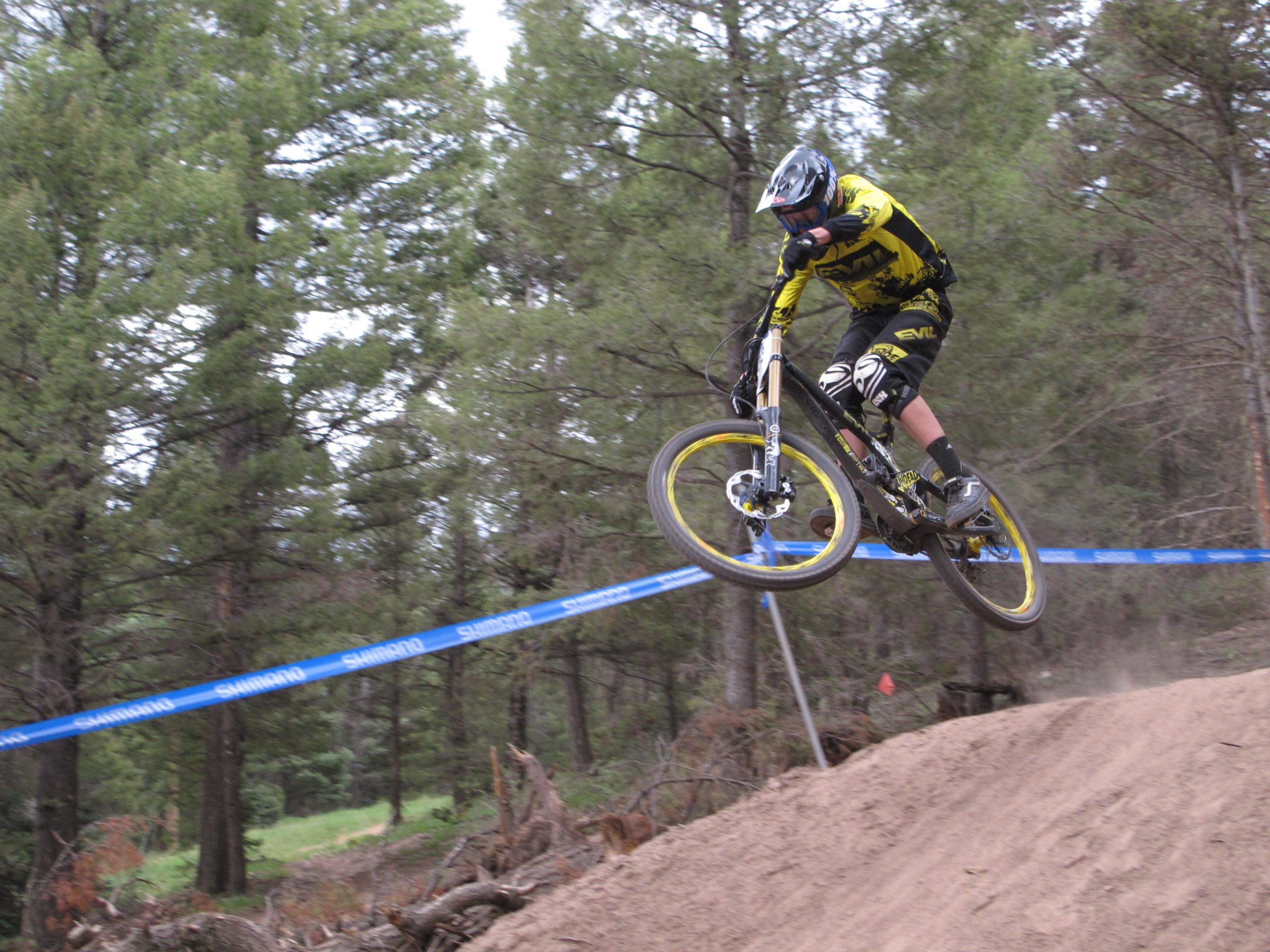 A mountain biker in yellow and black gear catches air while performing a jump on a dirt track surrounded by tall pine trees. A blue racing banner is visible in the background, suggesting a competitive setting. Dust is kicked up from the landing area, emphasizing the action of the moment. Angel Fire Bike Park mountain bike trail.