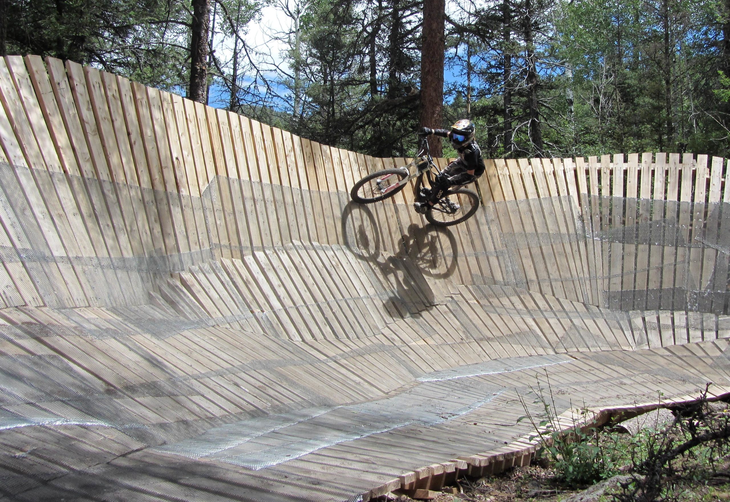A mountain biker performing a trick on a wooden pump track surrounded by trees, showcasing a dynamic motion as they ride along the curved, slatted surface. The scene is set in a natural outdoor environment with blue sky visible through the tree branches. Angel Fire Bike Park mountain bike trail.