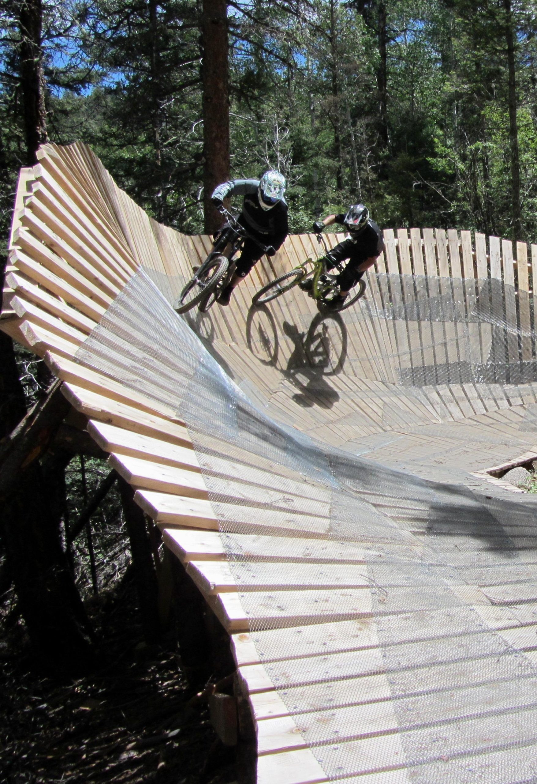 Two mountain bikers navigating a curved wooden track in a forested area, showcasing the thrill of downhill biking. The track features a wooden ramp with a mesh surface, surrounded by trees and a clear blue sky. Angel Fire Bike Park mountain bike trail.
