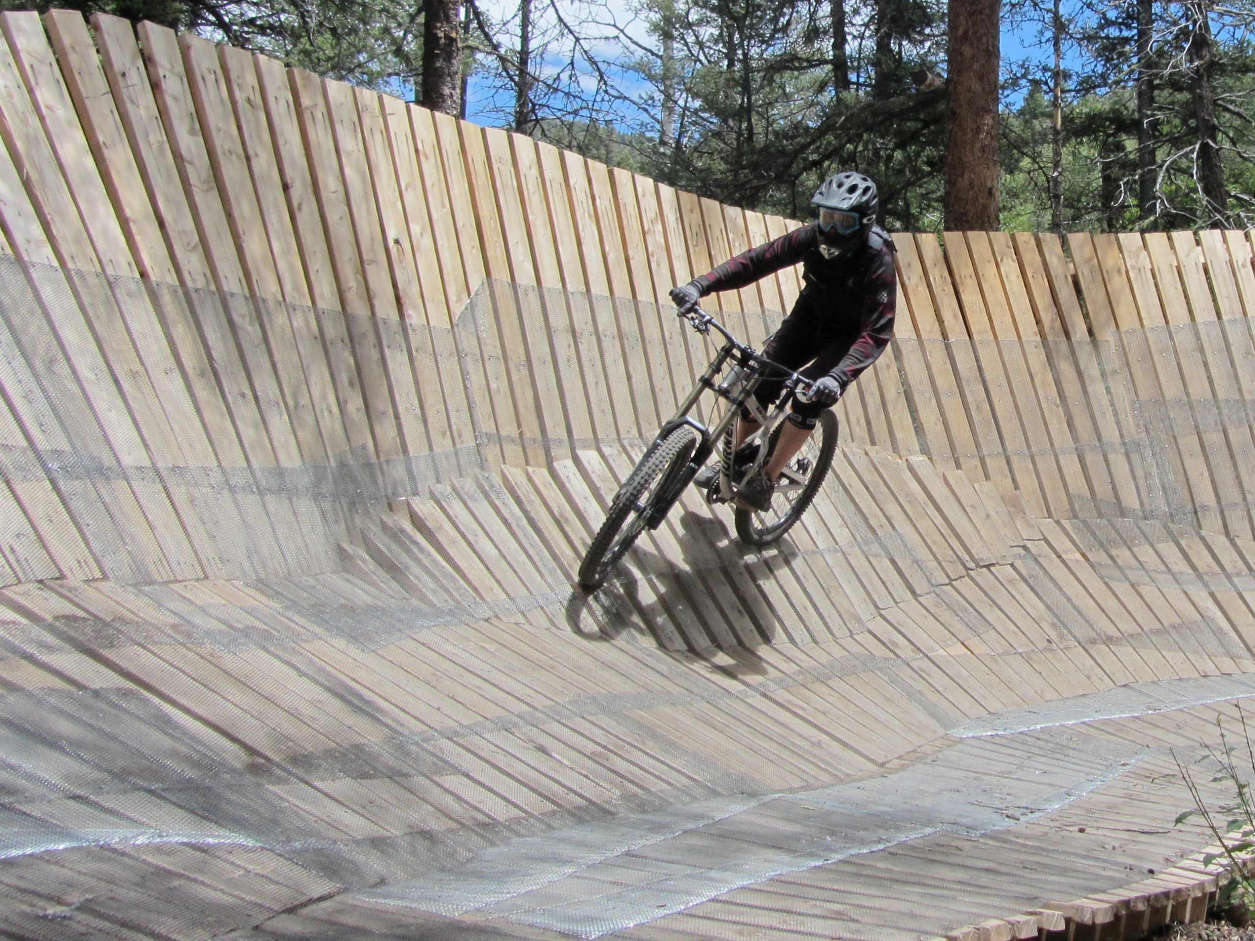 A mountain biker navigates a wooden pump track, leaning into a curve while wearing a helmet and protective gear. The track is surrounded by trees and features a series of undulating wooden surfaces designed for smooth riding. Angel Fire Bike Park mountain bike trail.
