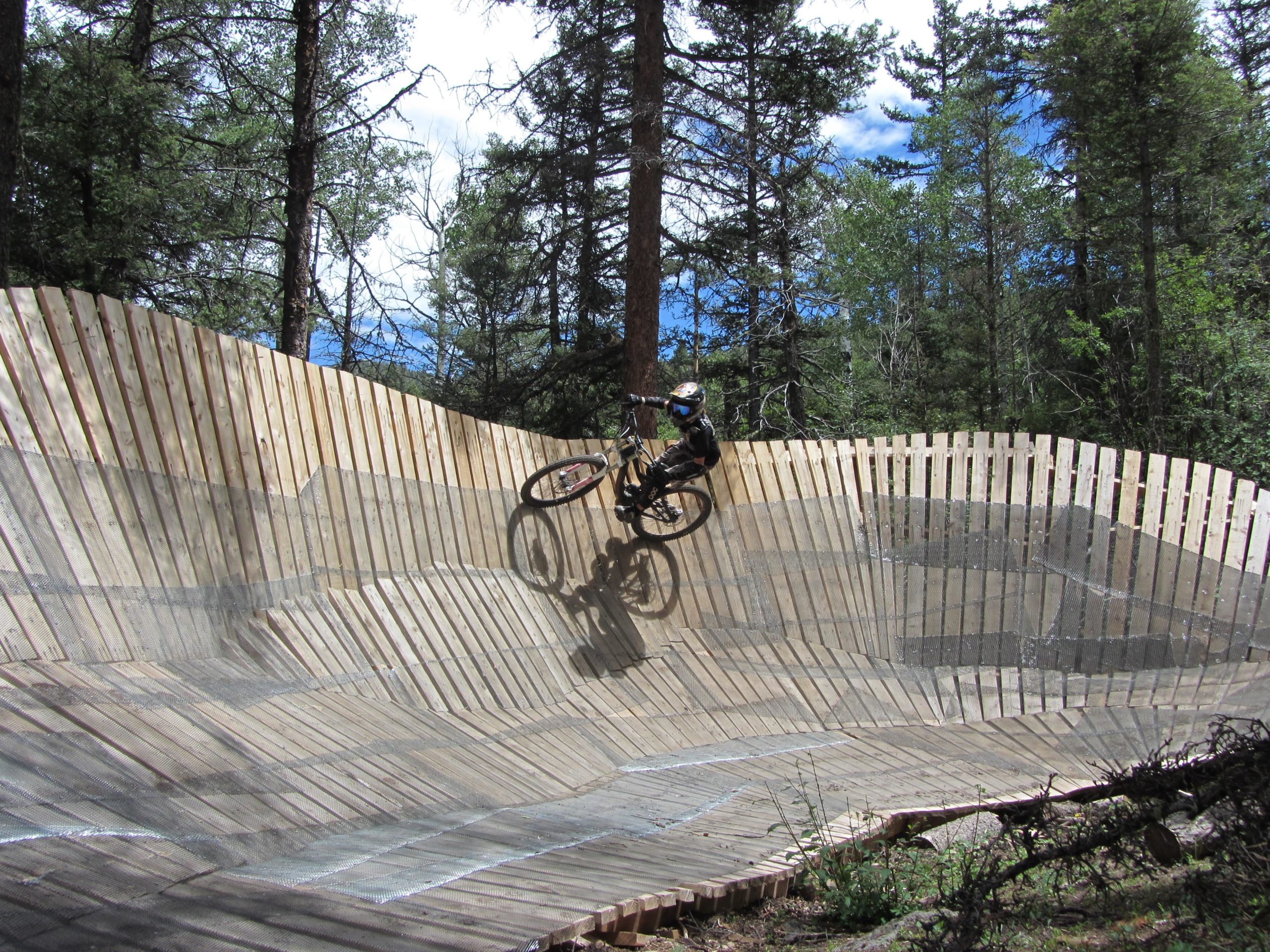 A mountain biker navigating a wooden pump track in a forested area, with tall trees in the background and a partly cloudy sky. The rider is leaning into a curve on the track, showcasing an energetic and dynamic stance. The distinctive wavy design of the wooden surface adds a unique element to the scene. Angel Fire Bike Park mountain bike trail.