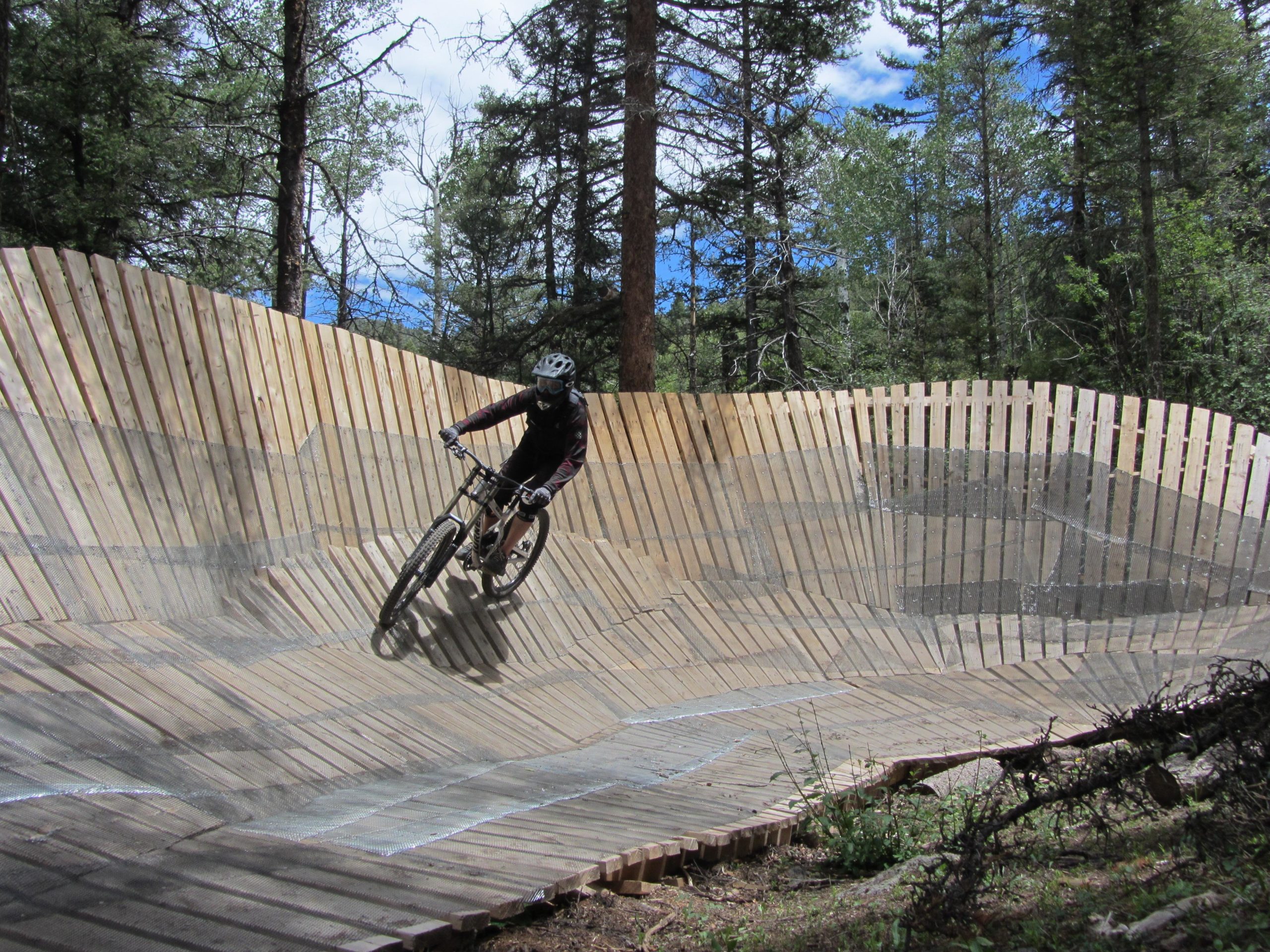A mountain biker expertly rides along a wooden pump track surrounded by tall trees. The track features a unique, wavy design, allowing for an exciting downhill ride. A clear blue sky peeks through the forest, highlighting the natural setting. Angel Fire Bike Park mountain bike trail.