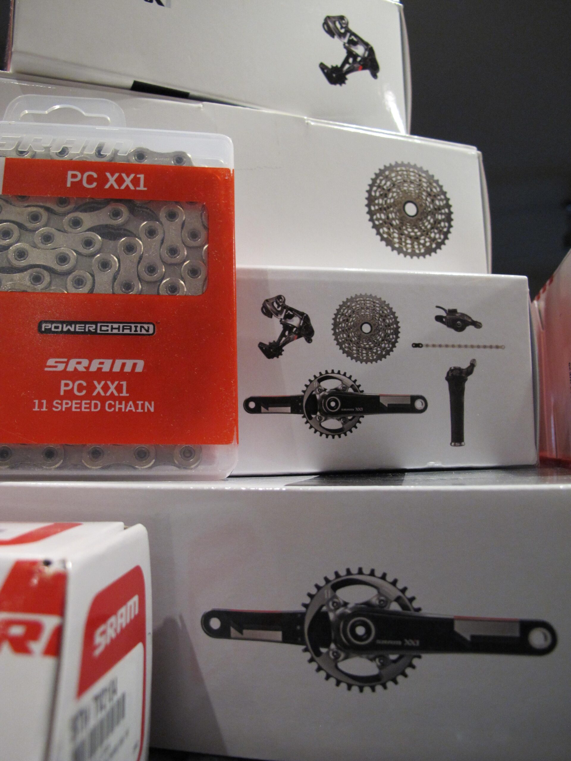 A close-up view of bicycle components from SRAM, including a package of the PC XX1 11 speed chain and various illustrated designs of chainrings and derailleurs on white boxes. The image highlights the details of the components, emphasizing their functionality for cycling enthusiasts.