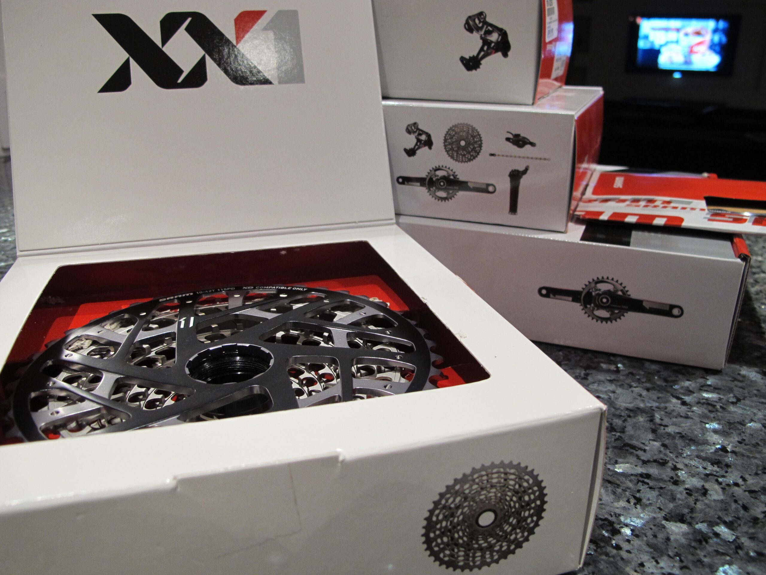 A close-up view of a bicycle component packaging, featuring a black and silver chainring prominently displayed inside a white box with the XX1 logo. Other boxes containing various bike parts are stacked behind it, with graphics illustrating their contents. The scene is set on a textured dark surface, and a television can be seen blurred in the background.