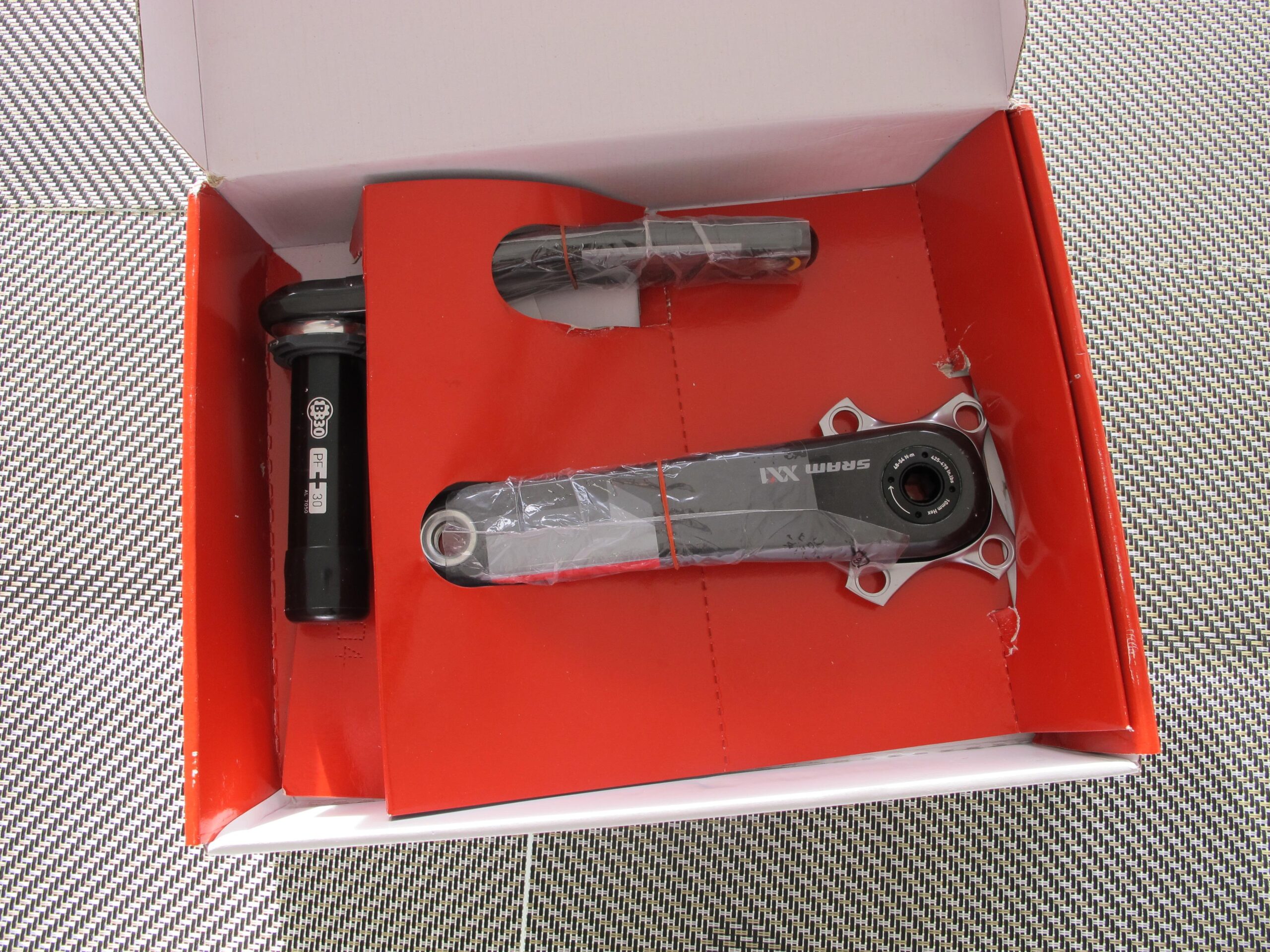 A close-up image of a SRAM XX1 mountain bike crankset and bottom bracket, displayed in a red cardboard box. The crankset is partially wrapped in plastic, showcasing its grey arms and black components. The bottom bracket is labeled with "PF30" and is also black. The background features a textured surface.