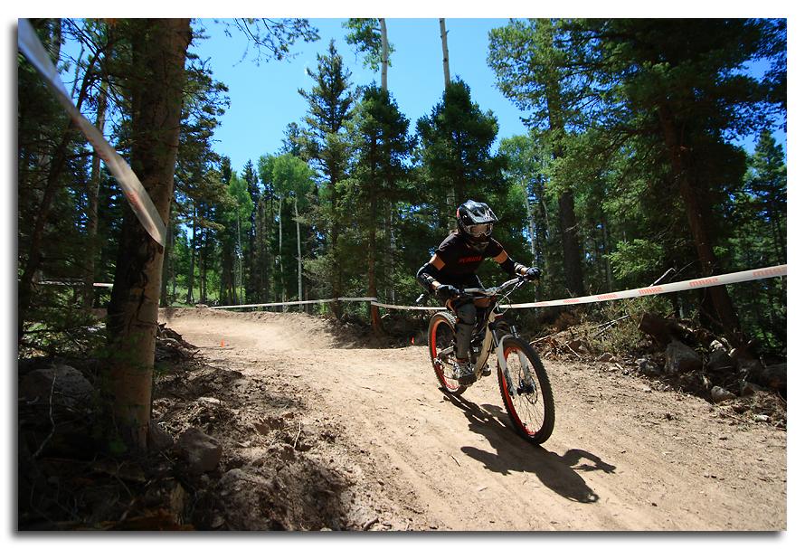 A mountain biker navigating a dirt trail in a forested area, surrounded by tall trees. The rider wears a helmet and protective gear, and the bike features bright orange accents. A course ribbon is visible alongside the path, indicating the trail boundaries. The scene is set on a sunny day with a clear blue sky. Angel Fire Bike Park mountain bike trail.